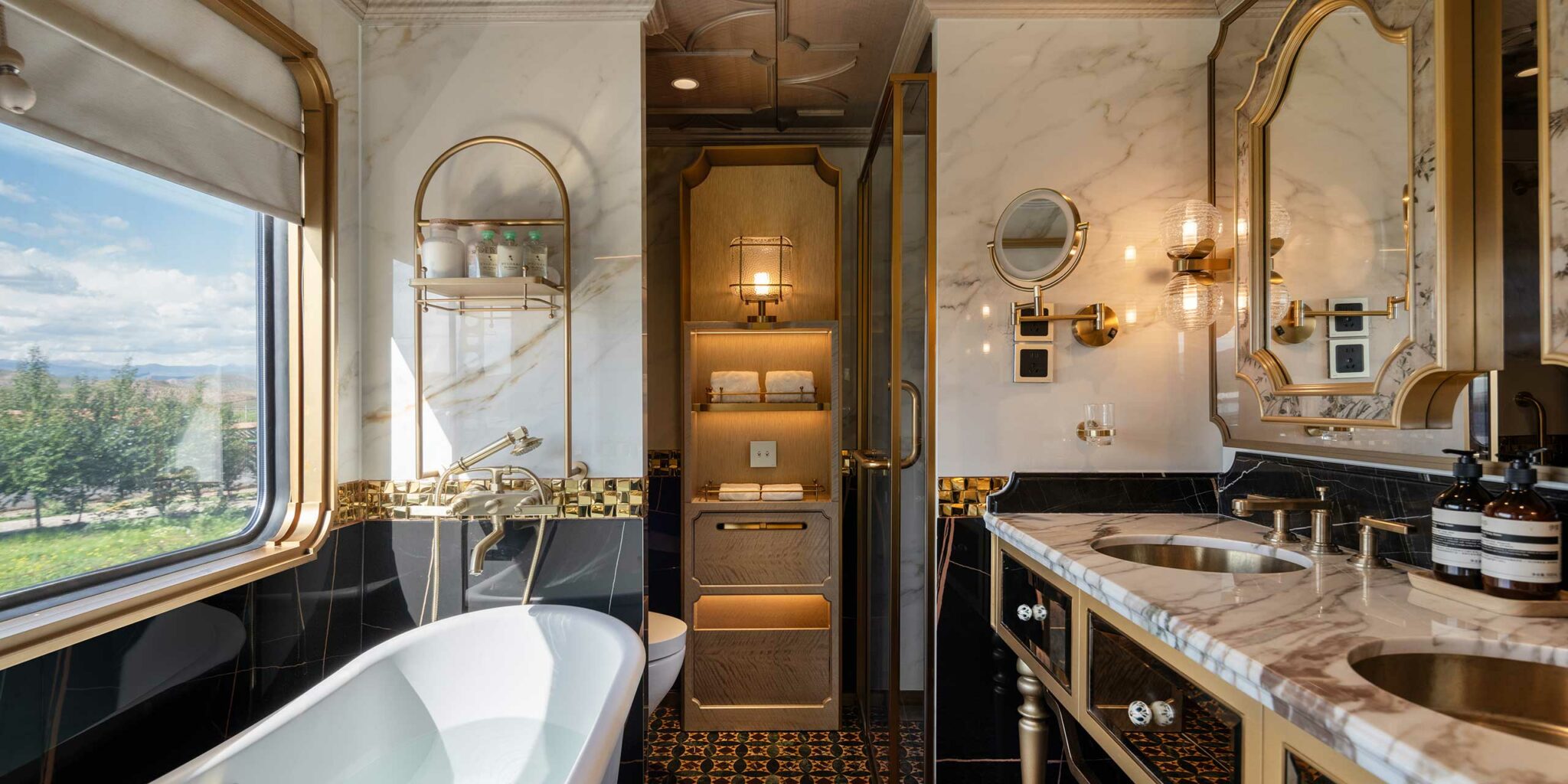 Luxurious train bathroom with elegant marble surfaces, gold fixtures, a freestanding tub, dual sinks, plush towels, and warm lighting. Spacious and opulent.