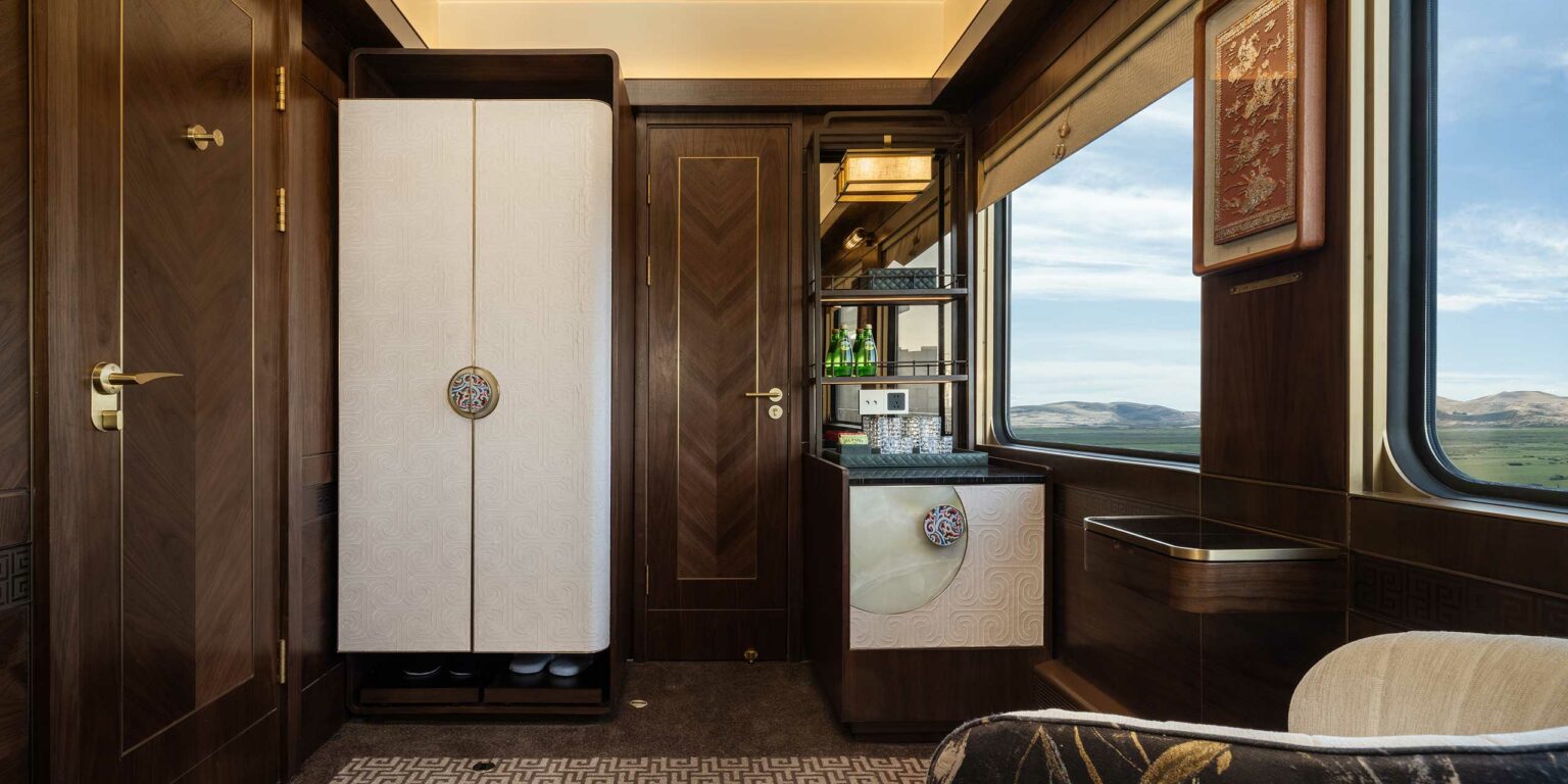 Luxurious train cabin interior with wood paneling and intricate designs. Features a wardrobe, small bar with green bottles, and a scenic window view.
