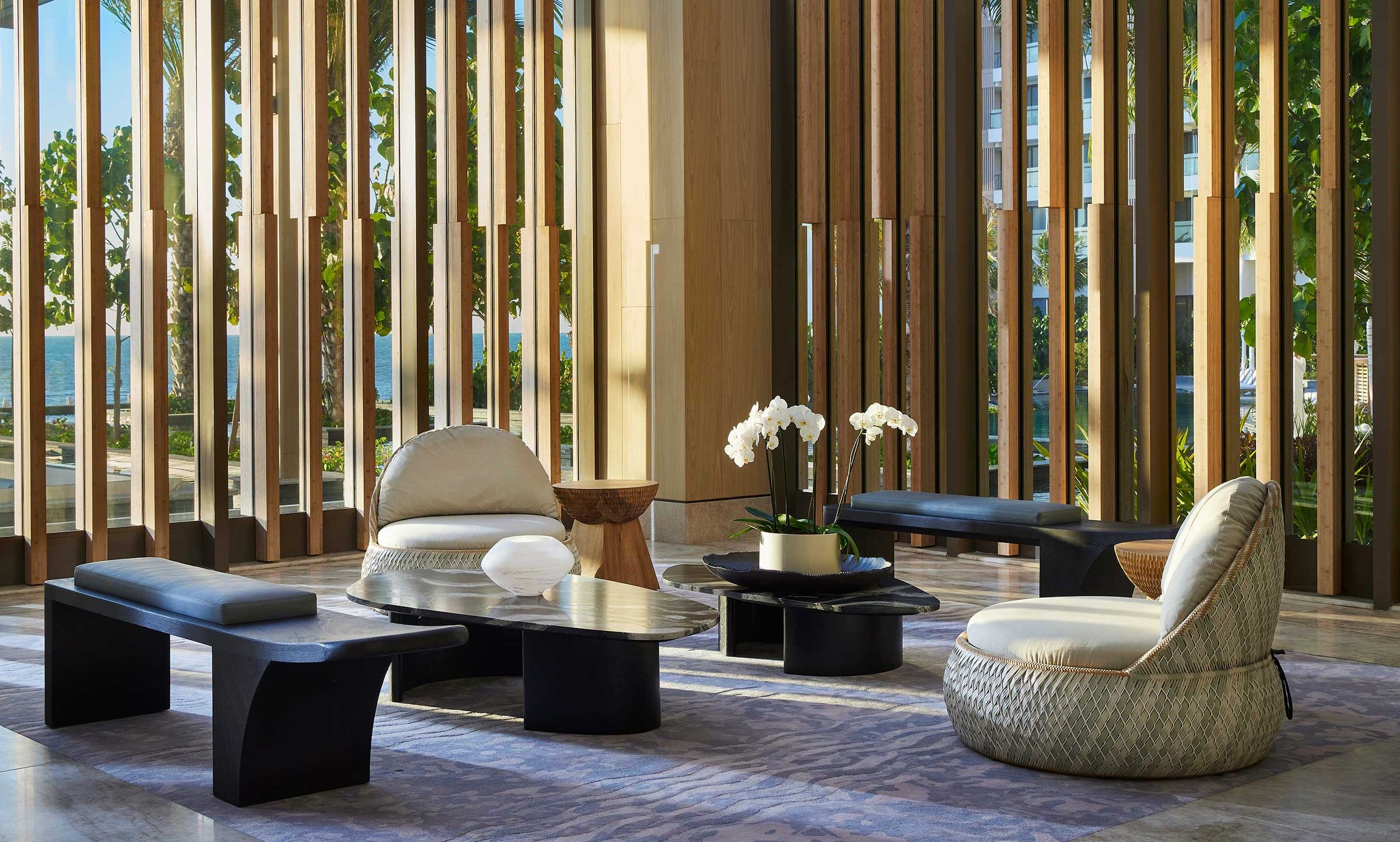 Spacious modern resort lounge with wood slat walls, minimalist black tables, cozy chairs, a potted orchid, and natural light creating a calm, elegant ambiance.