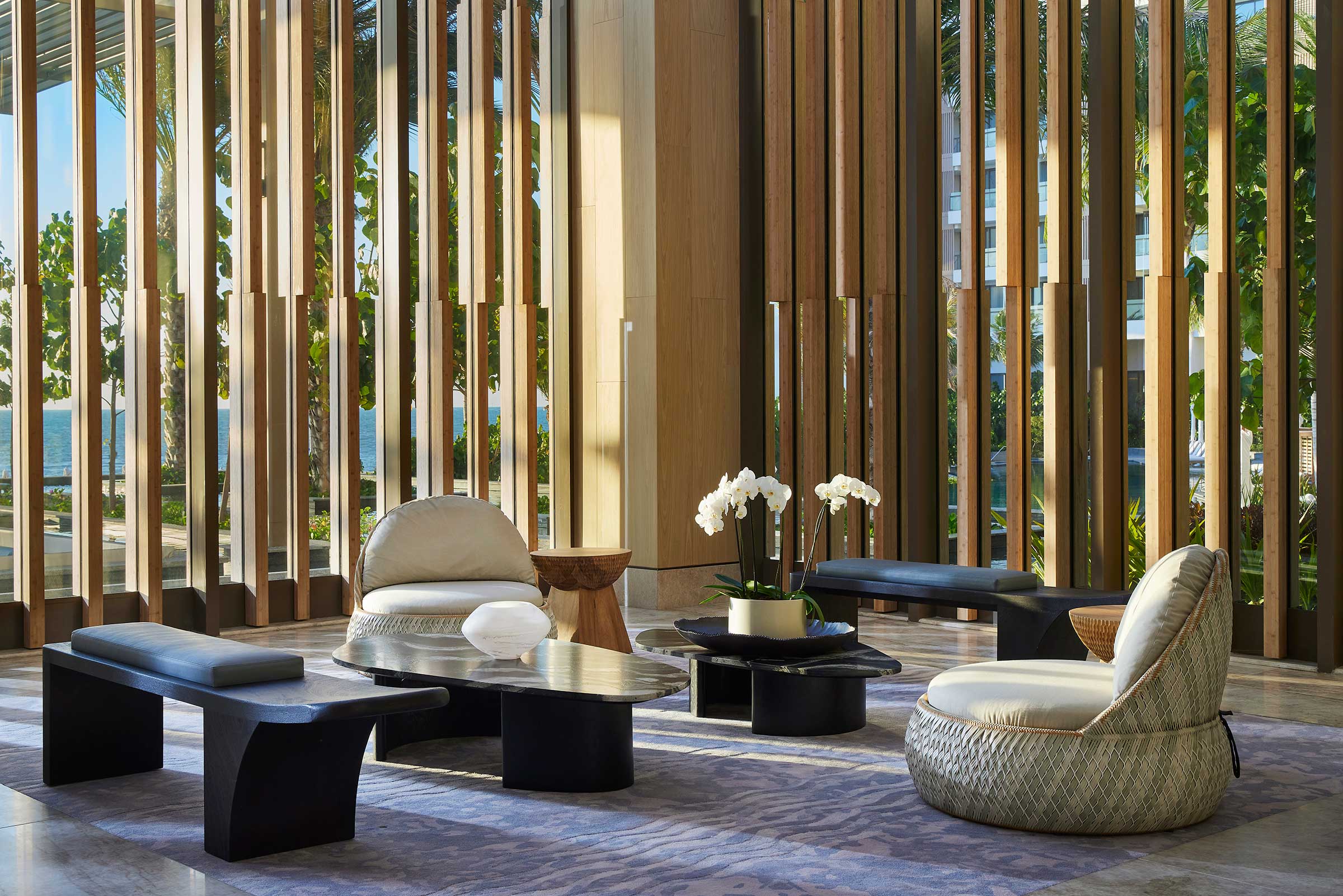 Spacious modern resort lounge with wood slat walls, minimalist black tables, cozy chairs, a potted orchid, and natural light creating a calm, elegant ambiance.