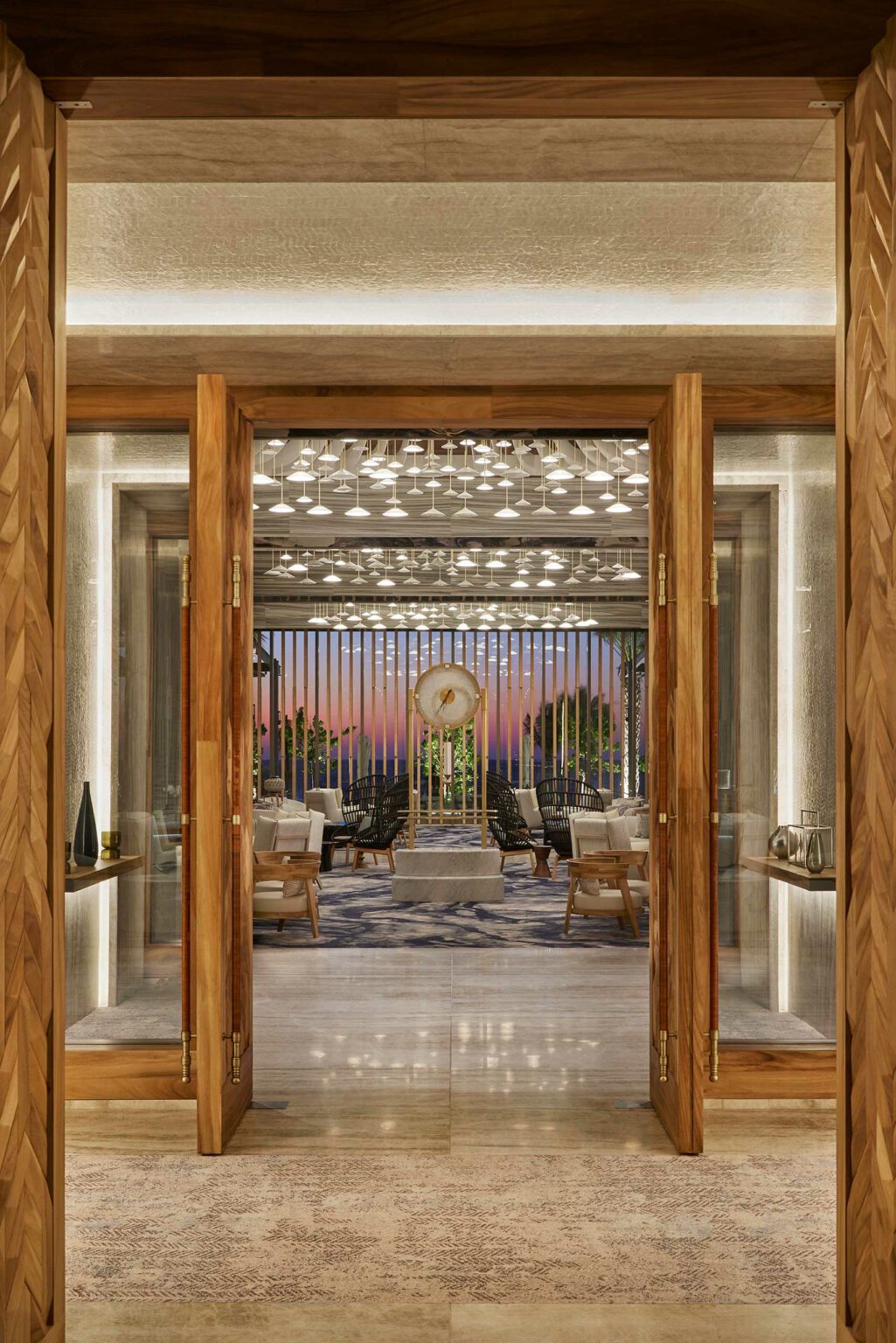 Elegant modern interior with wooden doors opening to a luxurious lounge. Features plush seating, a central sculpture, and sunset view through vertical blinds.