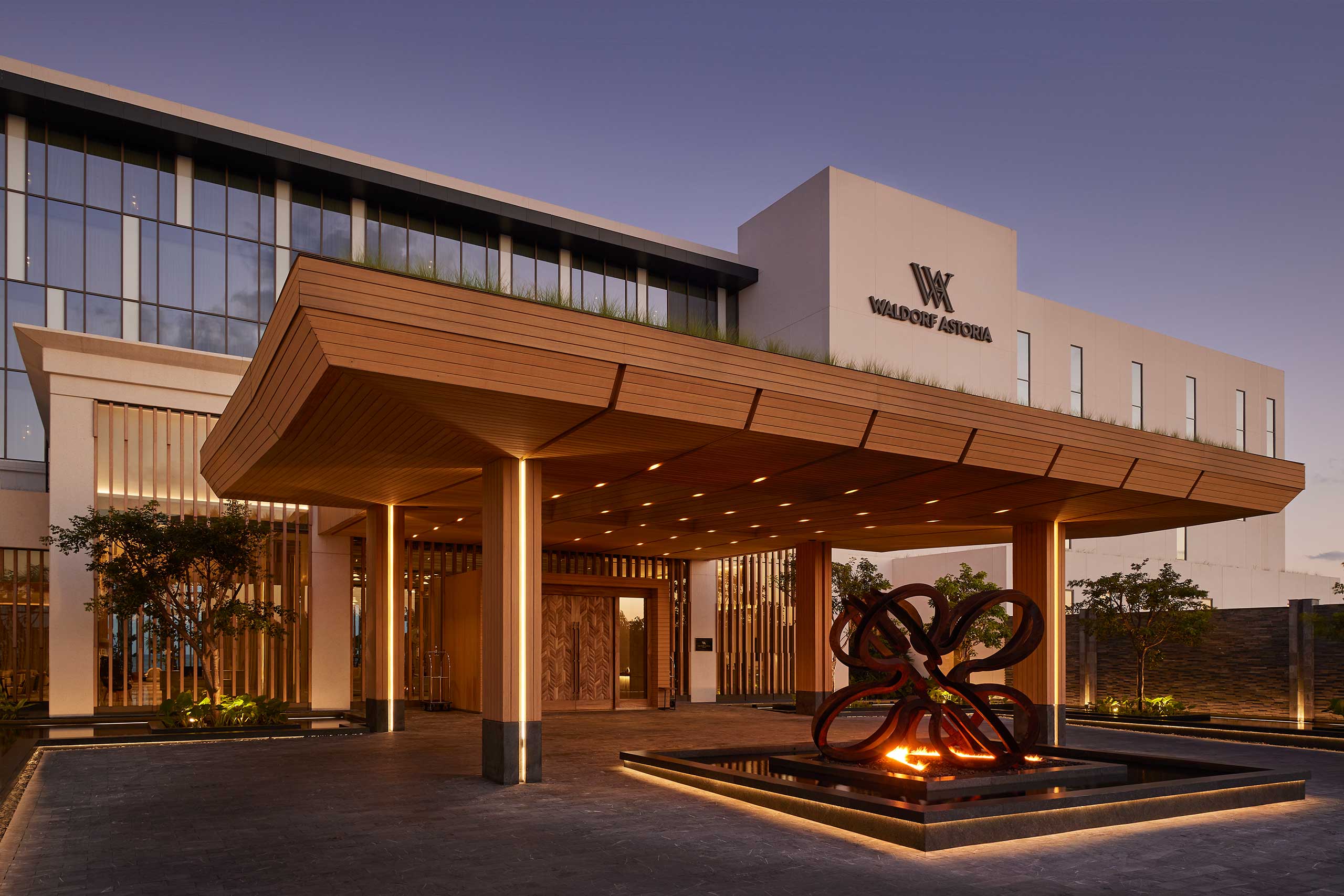 The image shows the entrance of Waldorf Astoria Riviera Maya resort featuring a wooden canopy and illuminated artwork in front, creating a warm and elegant ambiance at dusk.