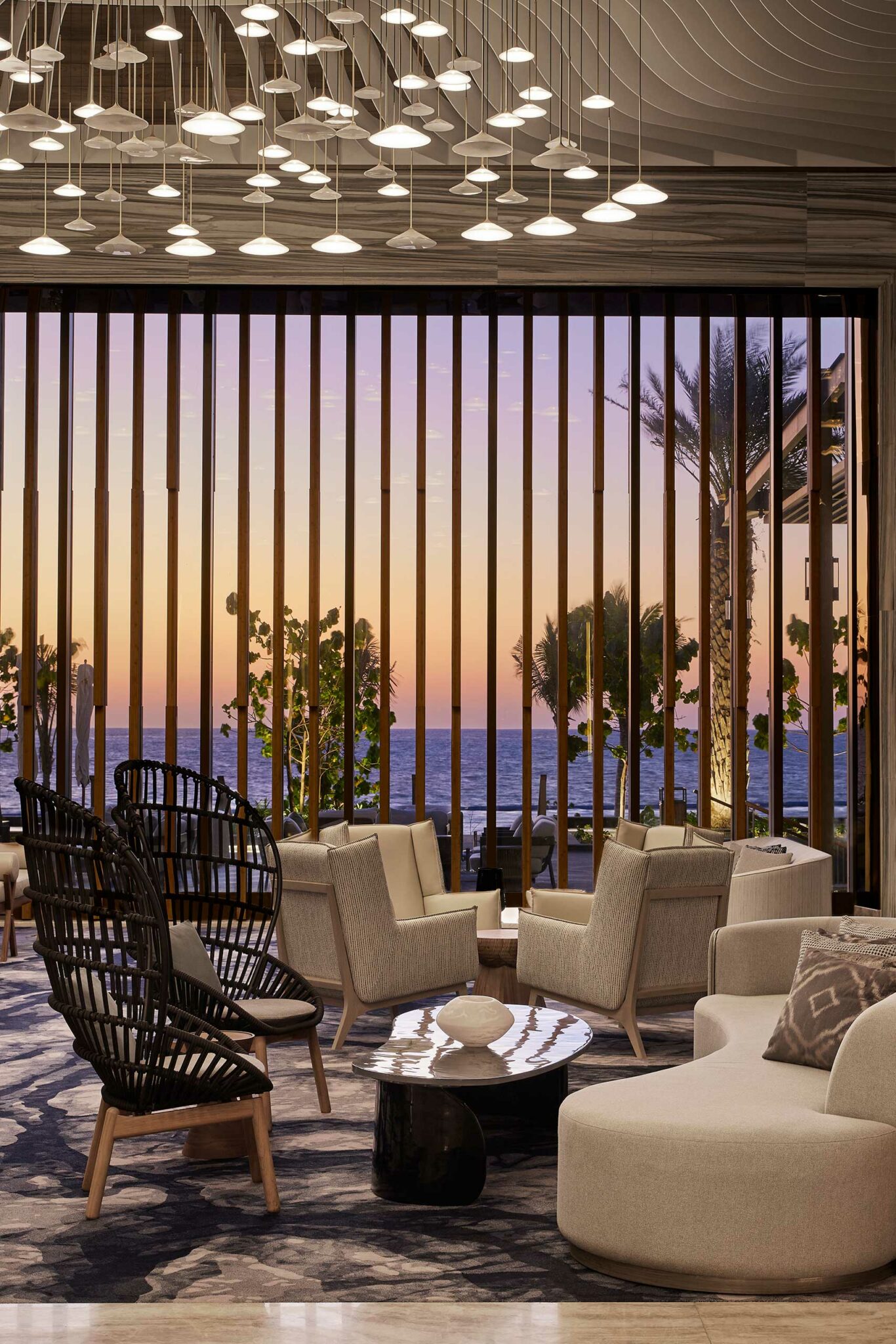 Modern resort lounge with wicker chairs and beige sofas facing a floor-to-ceiling window. Ocean and sunset view, creating a serene and luxurious ambiance.