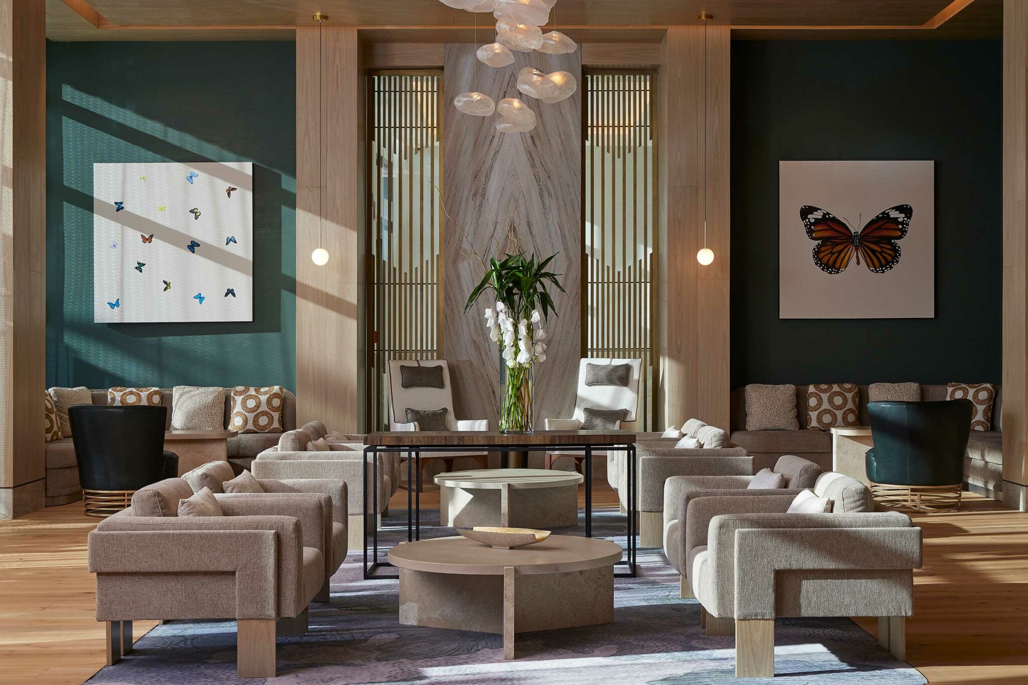 Elegant resort lounge with modern decor. Features gray seating, a central round table, butterfly artworks, a vase of flowers, and a sculptural ceiling light.