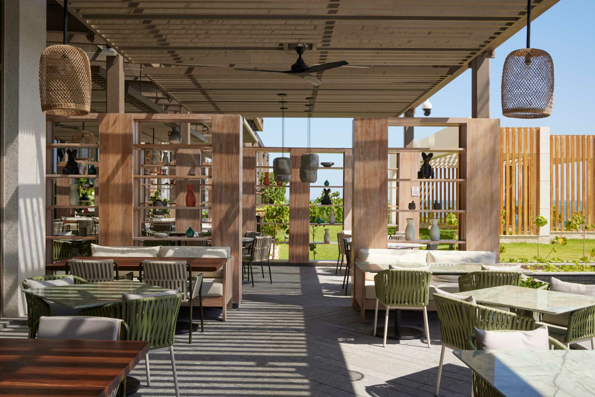 Resort outdoor dining area with modern decor, featuring wooden tables and chairs, wicker light fixtures, and open shelving. Sunlight casts shadows, creating a tranquil ambiance.