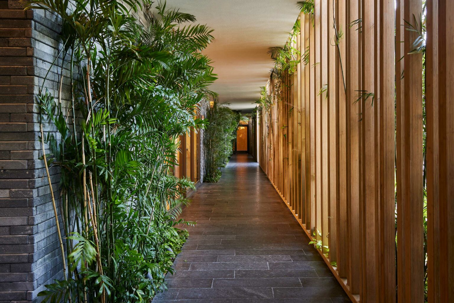 A serene resort hallway with lush green plants on the left, wooden slats on the right, and a stone floor, conveying a peaceful, natural atmosphere.