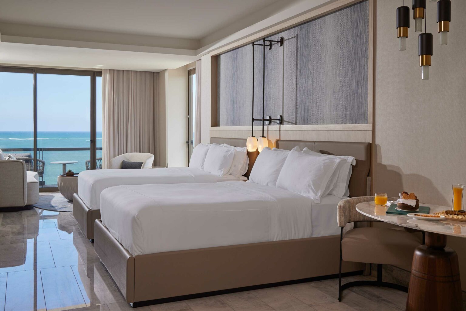 Hotel room with two neatly made beds, ocean view through large windows, soft lighting, and a small breakfast table with orange juice and pastries. Luxurious and serene atmosphere.