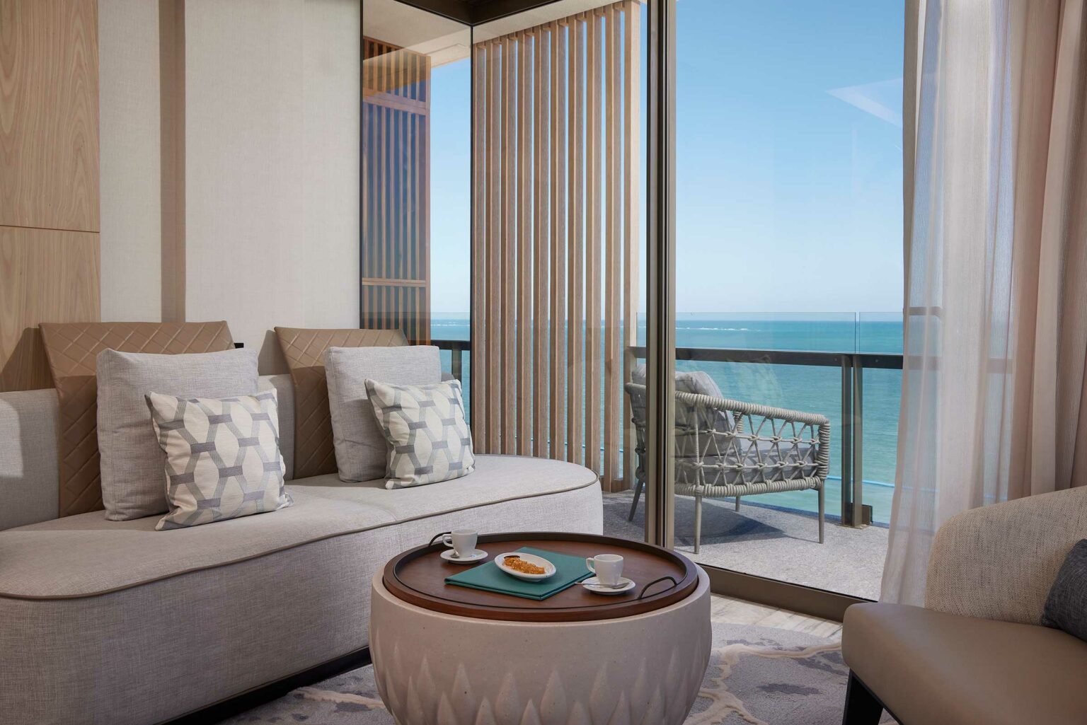 Contemporary suite living room with ocean view, featuring a beige sofa with patterned pillows, round coffee table with teacups and snack. Calm, inviting ambiance.