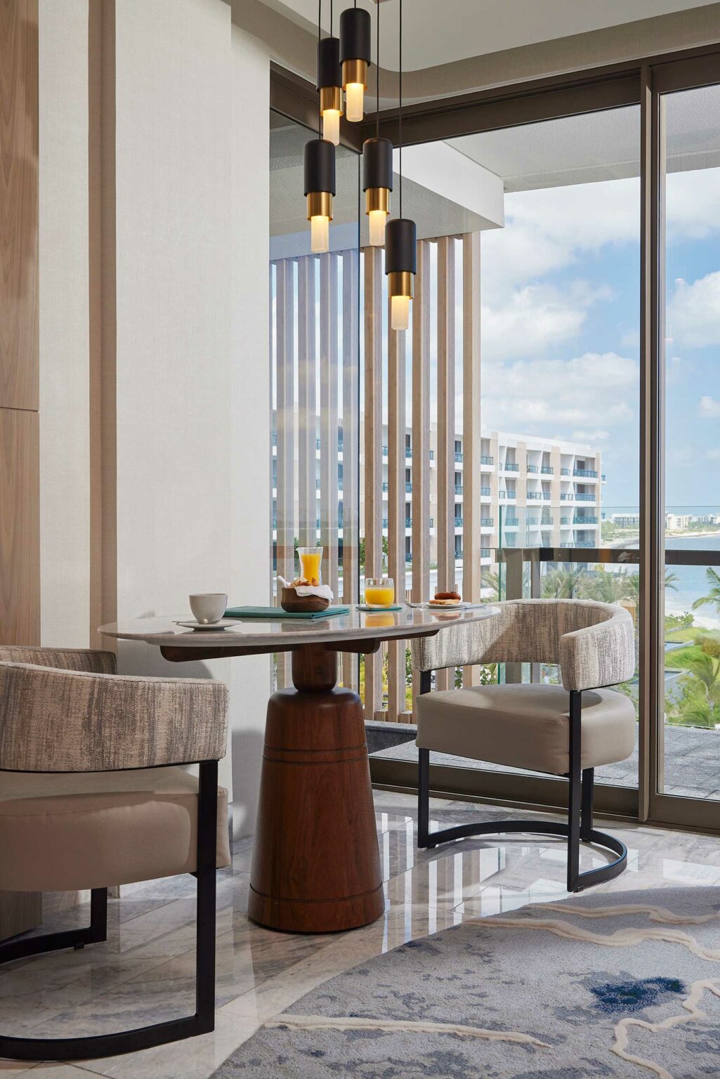 Modern resort dining nook with a round wooden table, two upholstered chairs, and pendant lighting, beside large windows overlooking a scenic view. Tablesetting includes juice, cups, and pastries, creating a cozy, welcoming atmosphere.