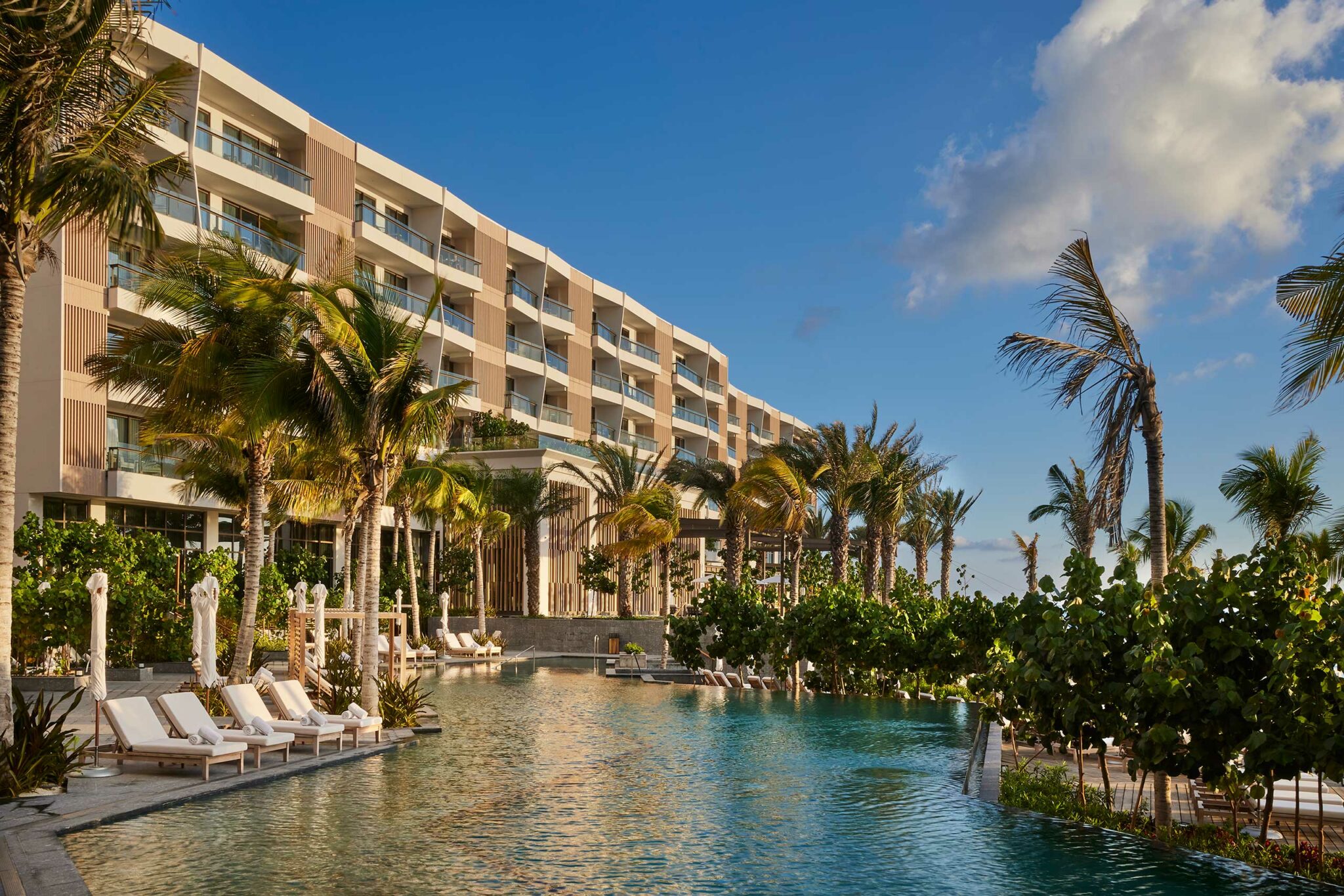 Modern beachfront resort with palm trees and lounge chairs beside a sparkling blue pool. The atmosphere is serene and inviting under a clear sky.