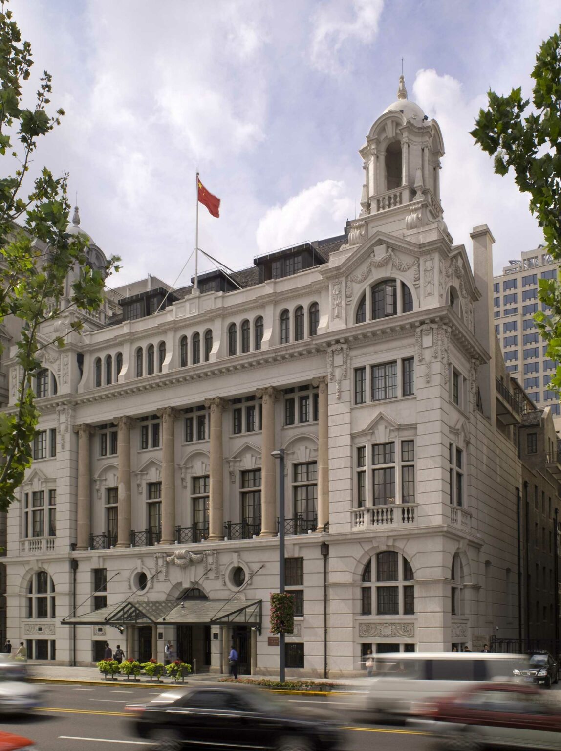 Exterior of Waldorf Astoria Shanghai on The Bund featuring a majestic white building with ornate architecture and columns, flying a red flag. Tall windows, busy street, and trees frame a bright, historic façade.