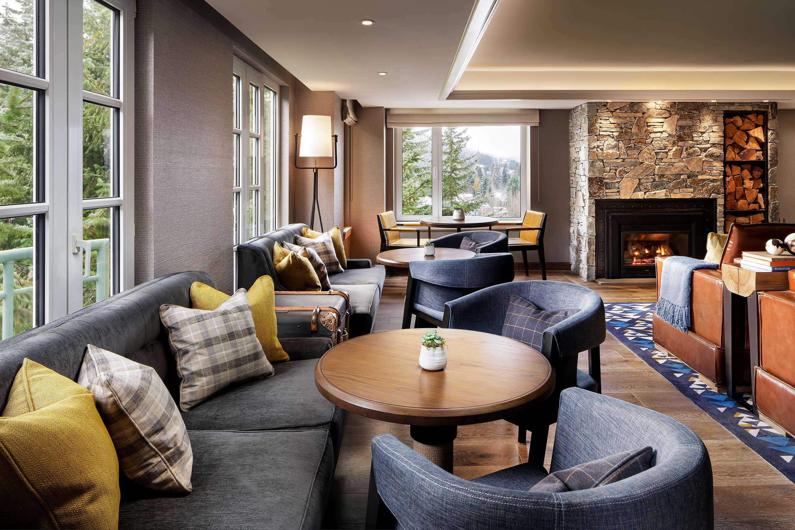 Elegant hotel lounge with gray sofas, yellow and plaid pillows, wooden tables, and blue chairs. Large windows offer forest views. Stone fireplace adds warmth.
