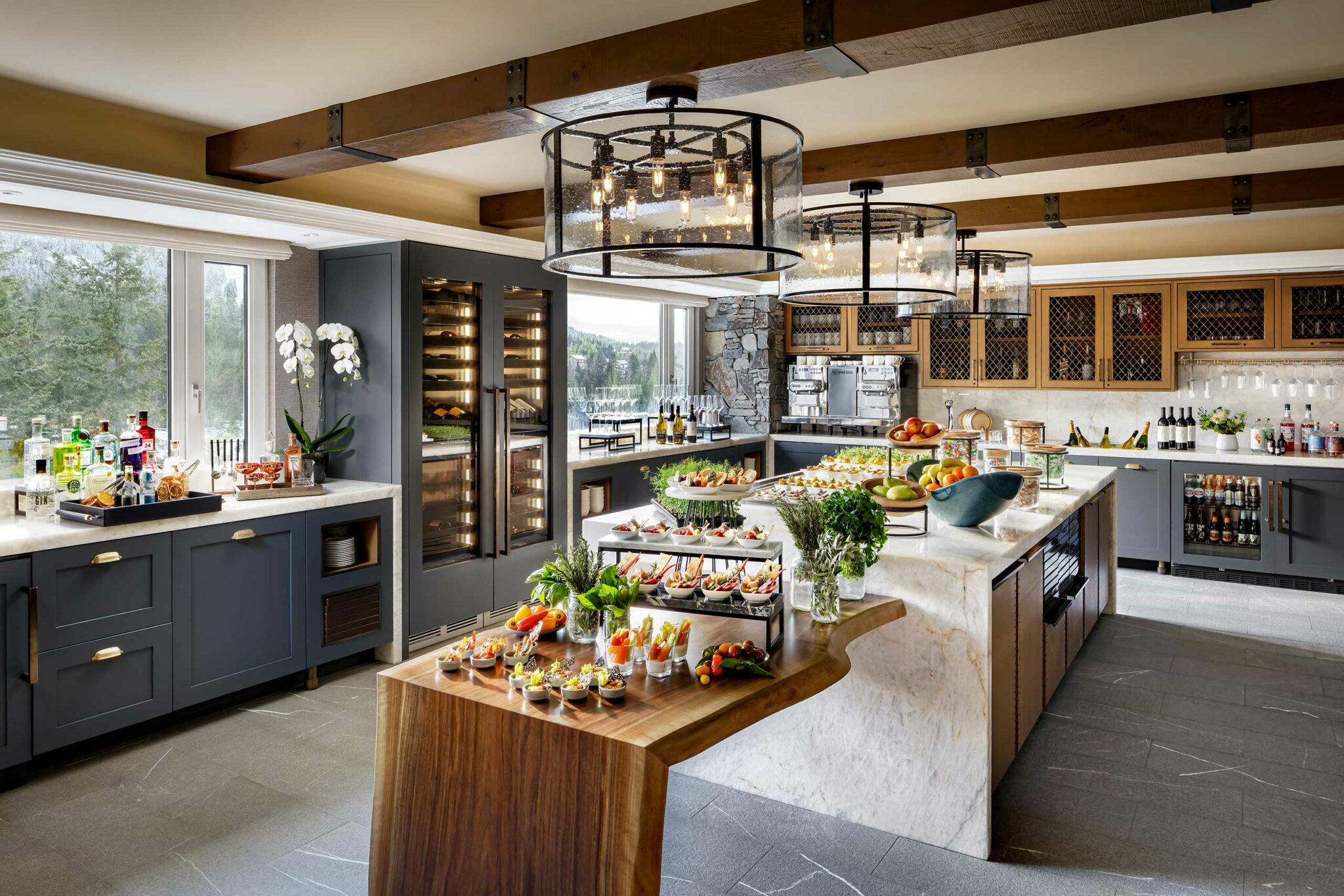 Luxurious hotel all day dining area with elegant chandeliers, marble island, hors d'oeuvres on wooden table, wine fridge, and large windows providing natural light.