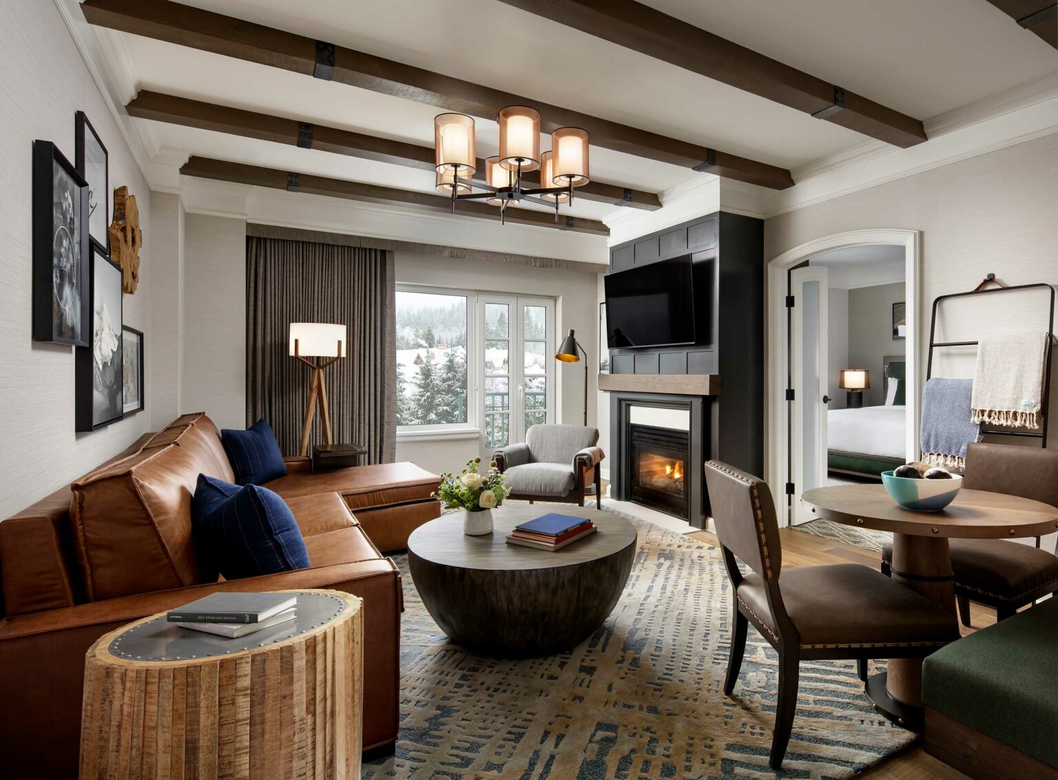 Elegant suite living room with a leather sofa, round wooden coffee table, and a fireplace beneath a mounted TV. Large windows show a snowy landscape.