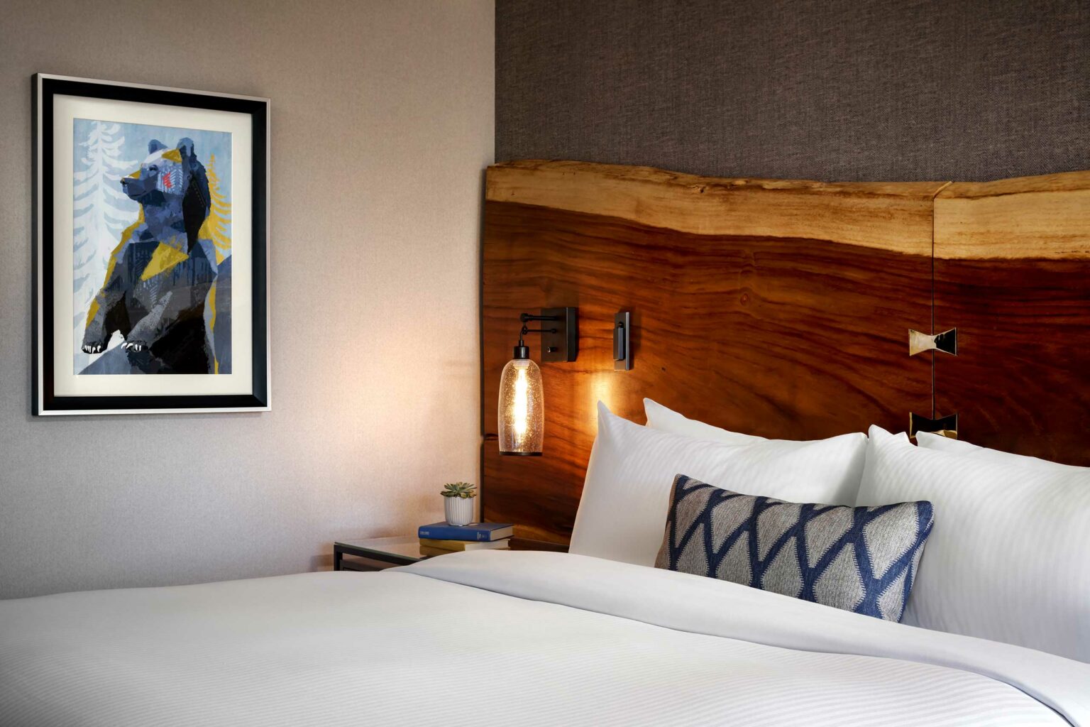 Contemporary hotel room with a natural wood headboard, crisp white bedding, and a geometric-patterned pillow. A colorful bear painting adorns the wall.