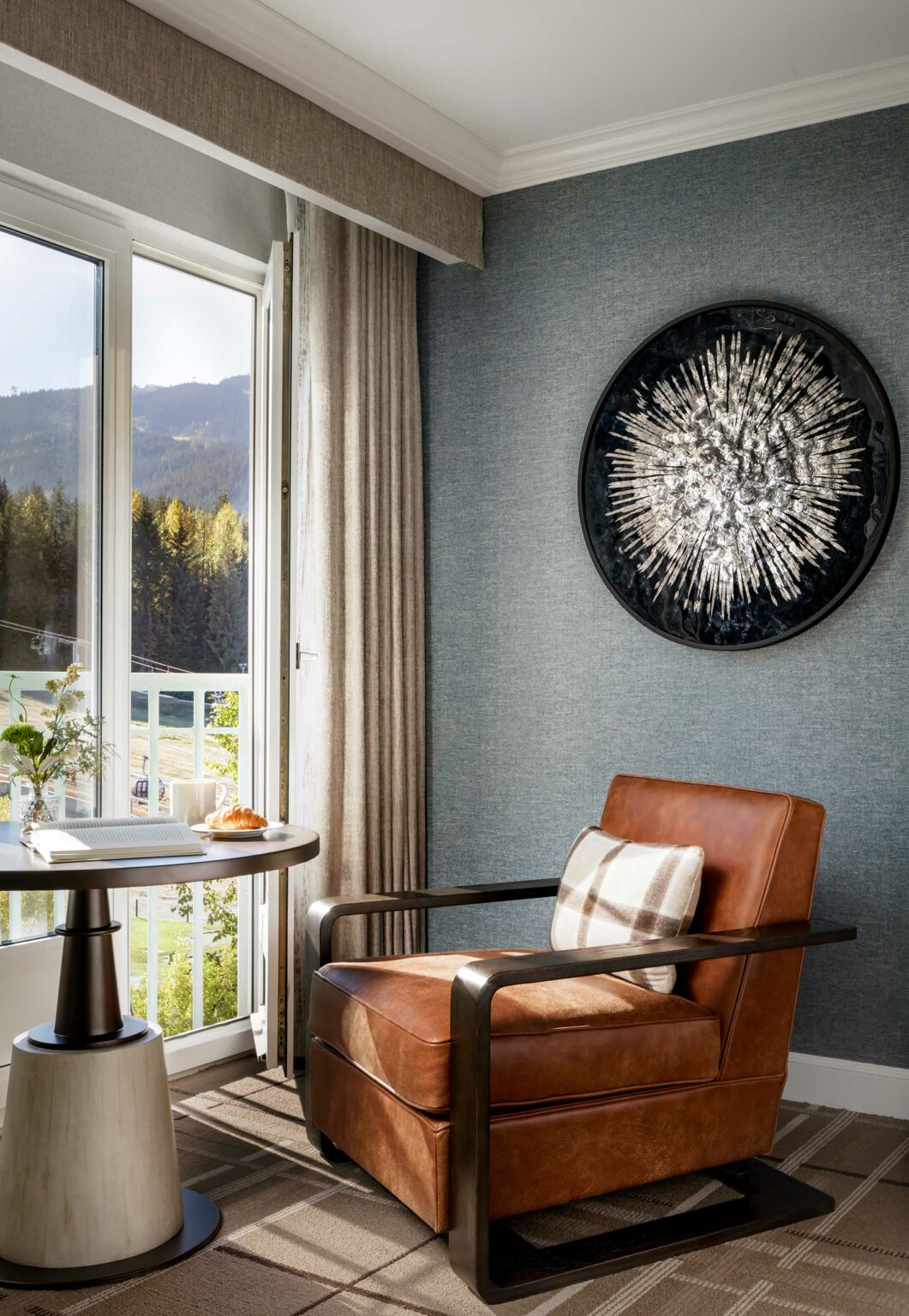 Cozy suite living room with a brown leather armchair beside a window overlooking mountains. A small table holds a croissant and book. Wall art adds elegance.