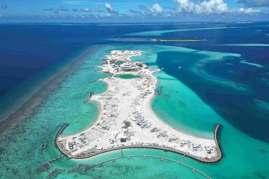 Aerial view of a bright, sandy island surrounded by turquoise and deep blue waters. Sparse buildings and construction are visible, with curved breakwaters extending into the sea. Fluffy clouds dot the clear blue sky, creating a serene, expansive atmosphere.