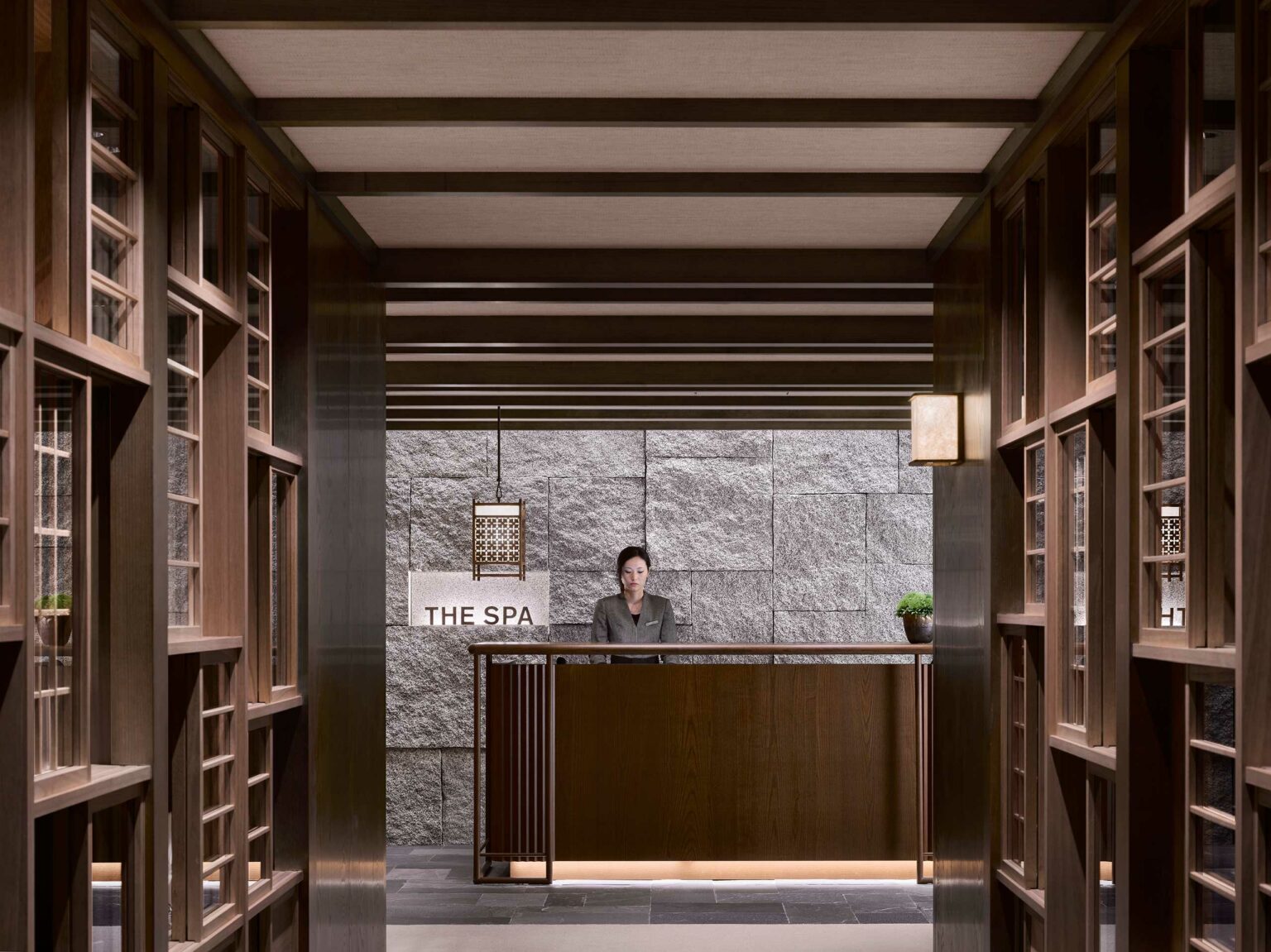 Stylish hotel spa lobby with wooden decor, featuring a reception desk with a person behind it. Soft lighting and a calming ambiance set a tranquil tone.