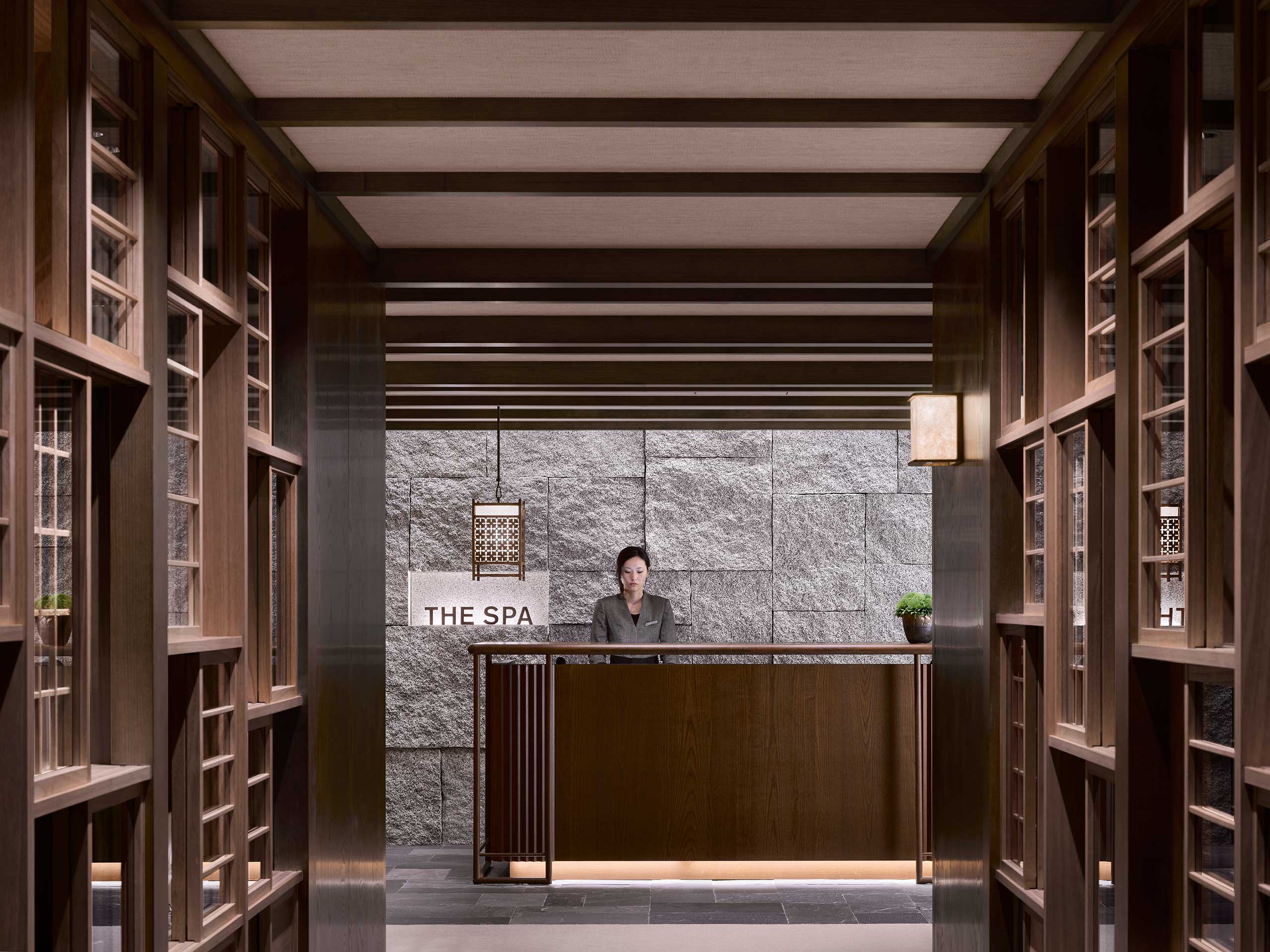 Stylish hotel spa lobby with wooden decor, featuring a reception desk with a person behind it. Soft lighting and a calming ambiance set a tranquil tone.