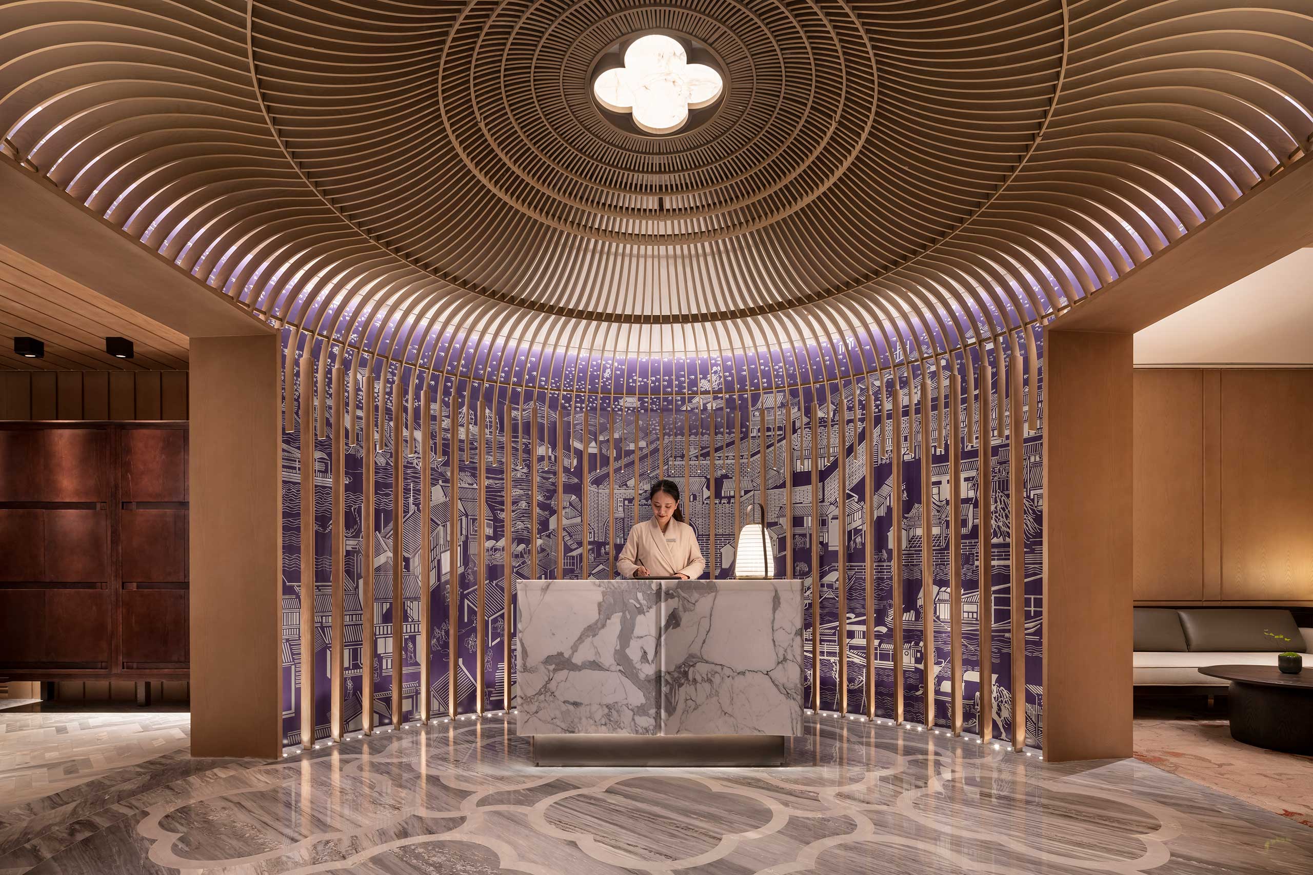 Luxurious hotel spa lobby with a marble reception desk. A receptionist stands beneath a dome-like ceiling with wooden slats and intricate wall art. Warm lighting sets a serene tone.