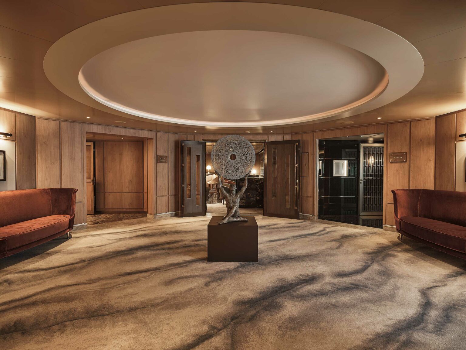 Spacious cruise lobby with warm lighting, features a circular ceiling design and a central abstract sculpture. Two maroon couches line the wood-paneled walls.