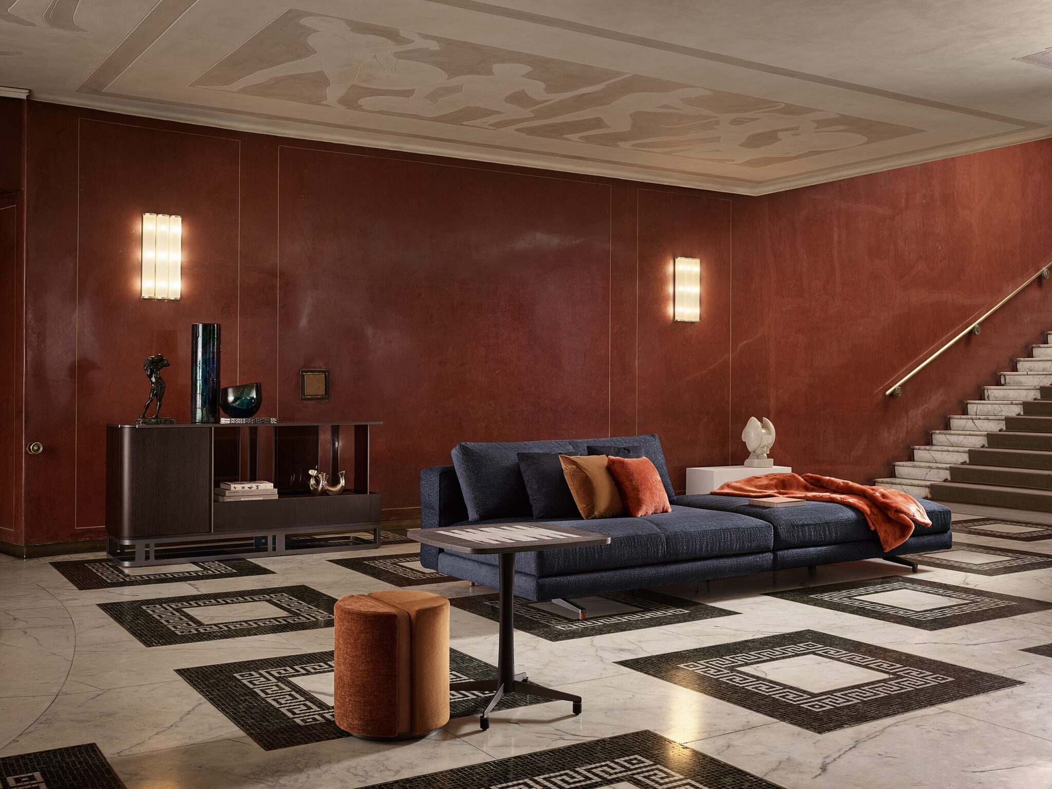 Luxurious living room with red walls, a sleek modern sideboard with dark wood finish, a navy sofa adorned with orange pillows and a throw, elegant lighting, and marble stairs. Sophisticated and warm.