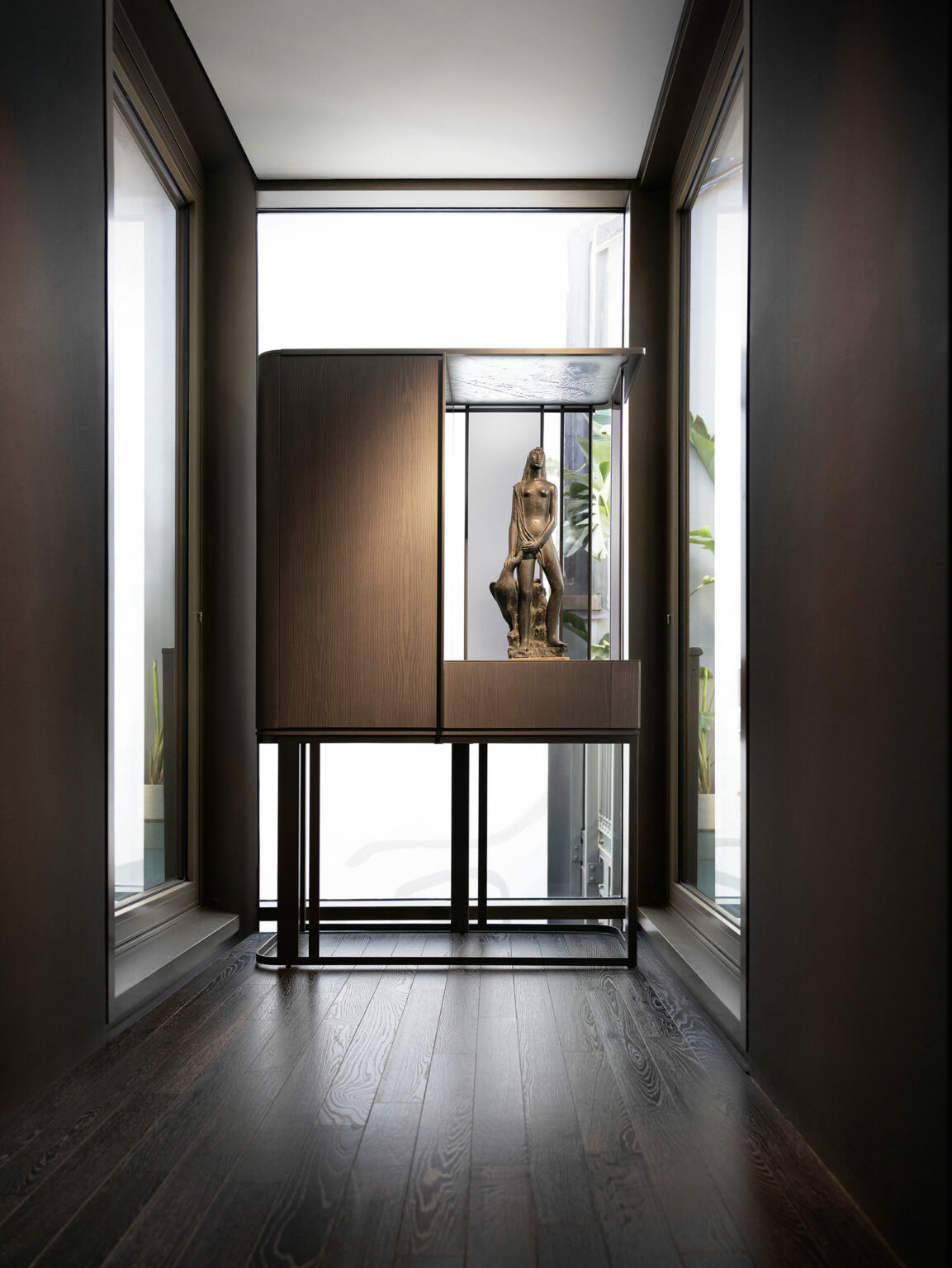 Elegant hallway with dark wood floors, flanked by large windows. A bronze statue is displayed in a modern brown cabinet on a minimalist metal frame with sleek vertical glass panels, creating a serene ambiance.