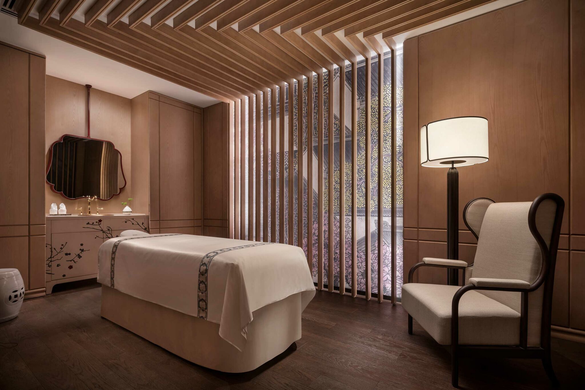 A serene hotel spa treatment room with a massage table covered in a white sheet, warm wooden paneling, decorative slats, a floral cabinet, and a soothing lamp.