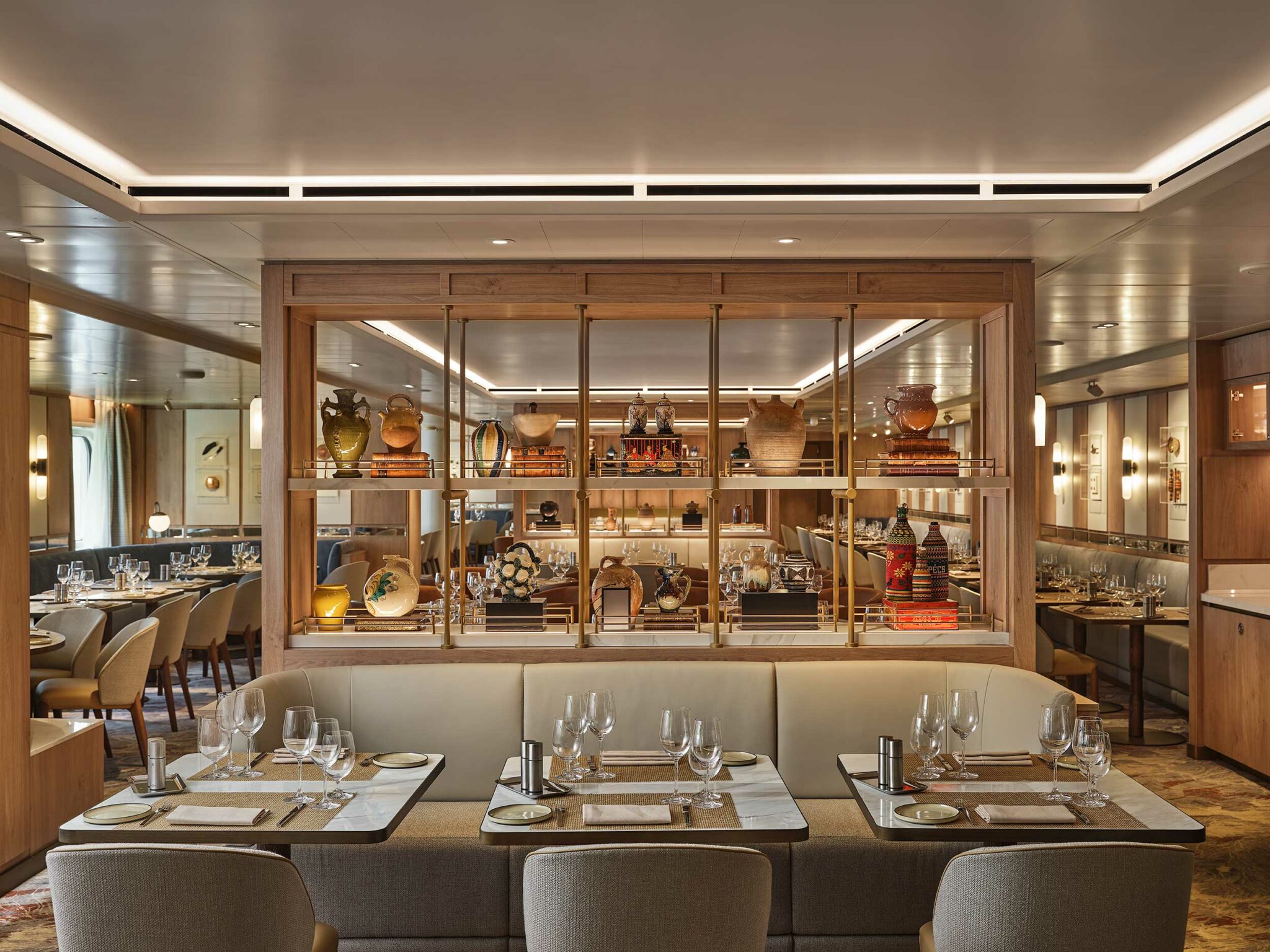 Elegant cruise restaurant interior with neatly set tables and plush seating. Wooden shelves displaying ornate vases create a cozy, sophisticated ambiance.