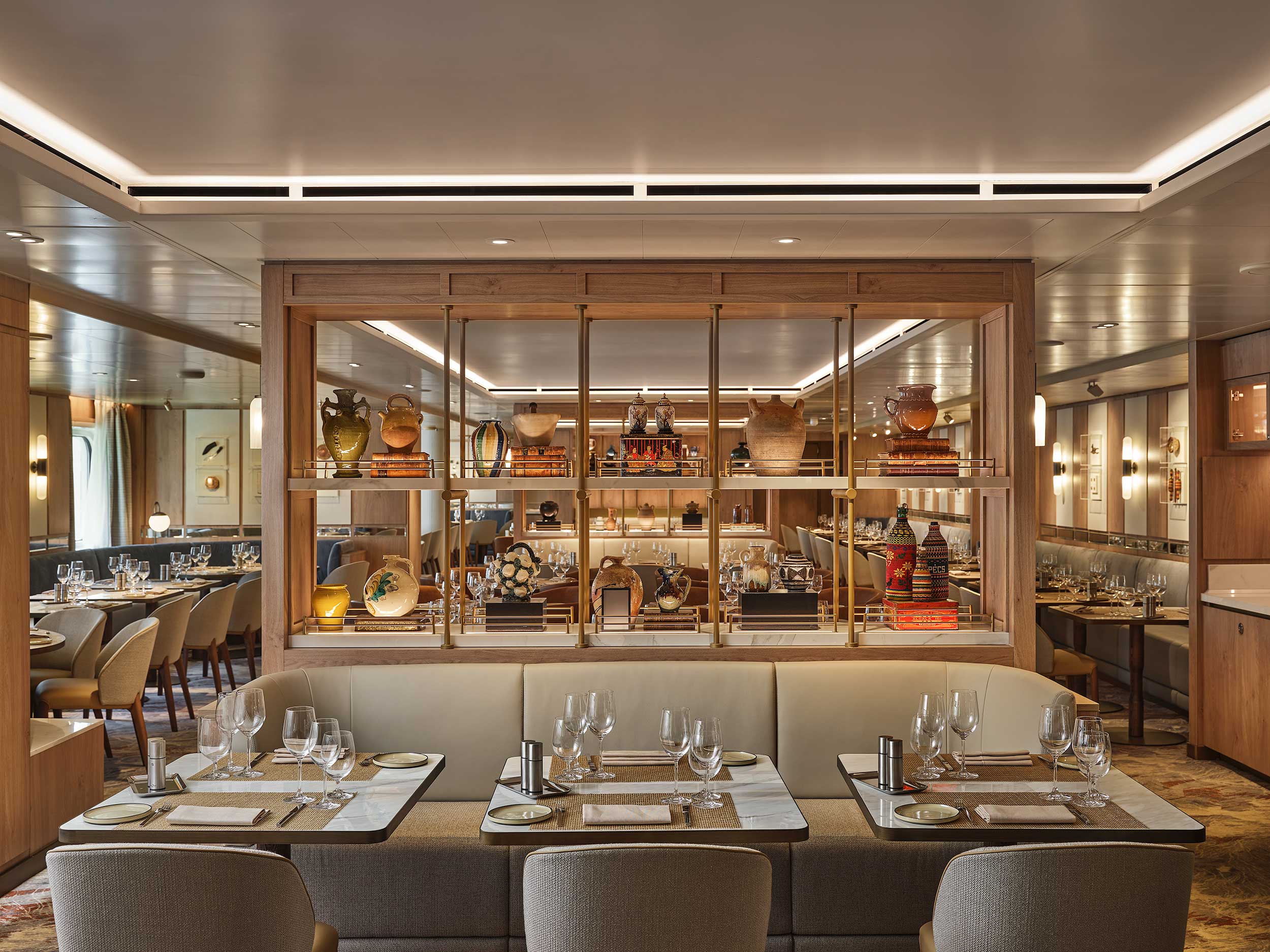 Elegant cruise restaurant interior with neatly set tables and plush seating. Wooden shelves displaying ornate vases create a cozy, sophisticated ambiance.