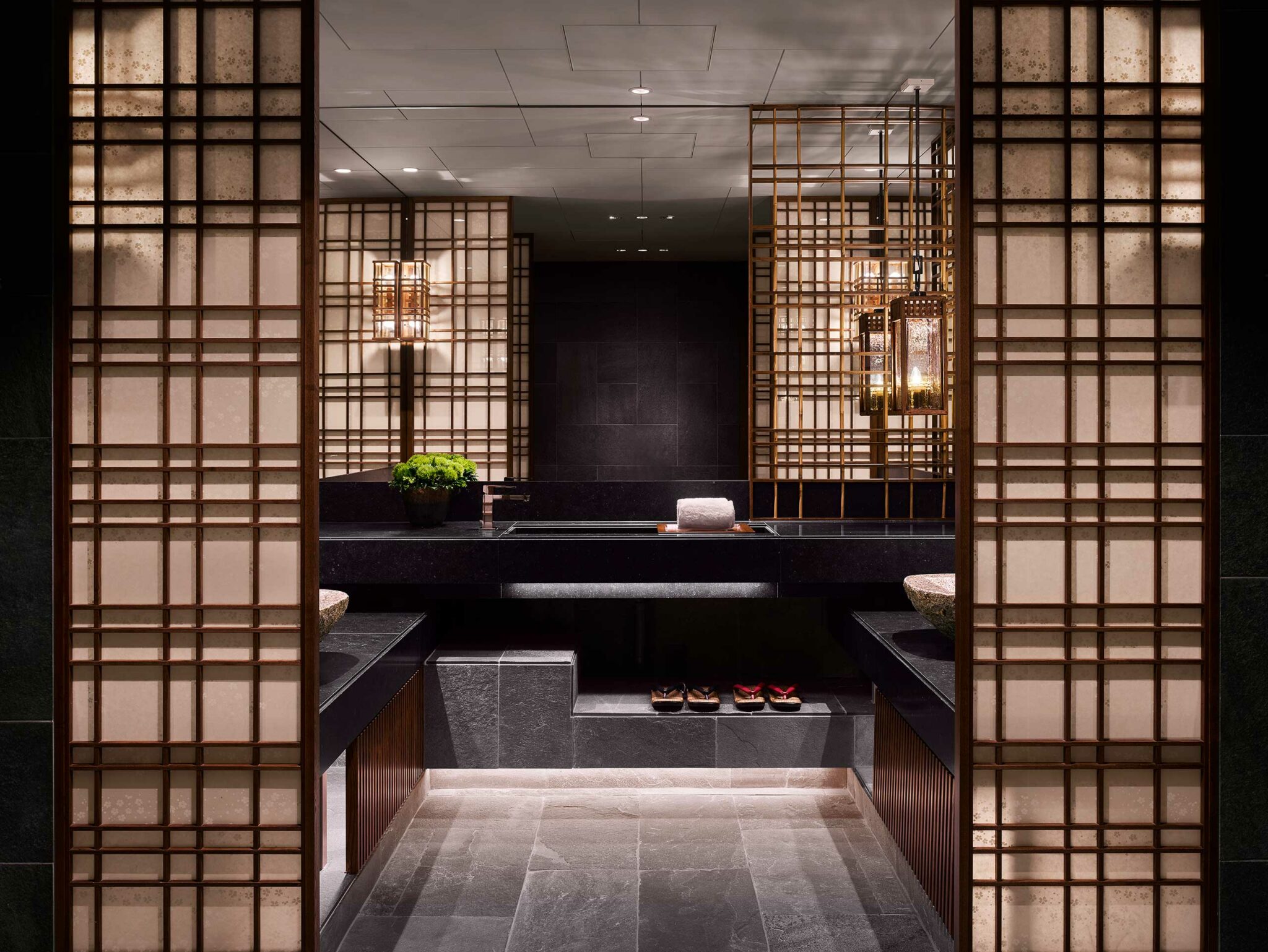 Luxurious hotel bathroom with dark stone surfaces, wooden lattice panels, warm lighting, a potted plant, folded towel, and neatly arranged slippers, exuding elegance.