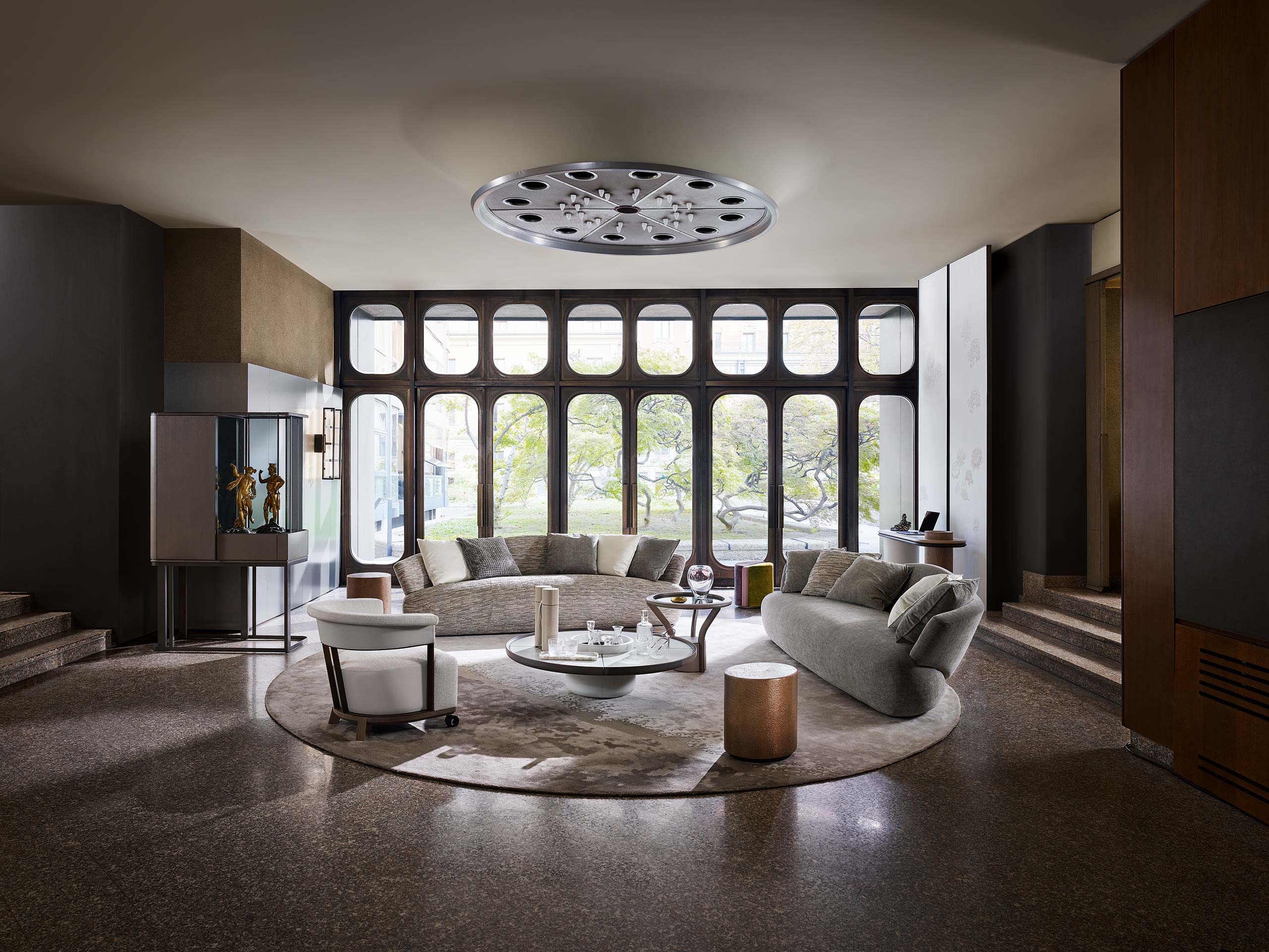 Spacious living room with large windows showing trees outside. Features a curved sofa, modern chairs, a round table, and a modern brown cabinet on a minimalist metal frame with sleek vertical glass panels. The soft lighting creates a serene ambiance.