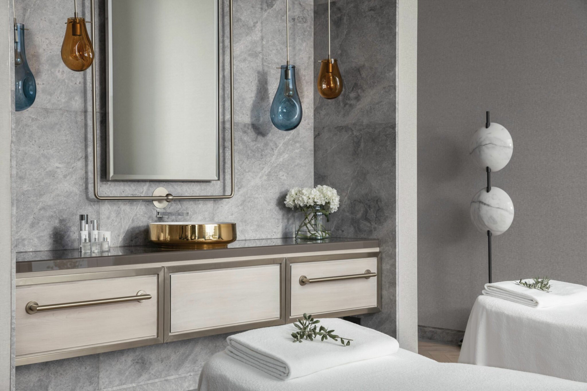 Luxurious spa treatment room with grey marble walls, modern pendant lights, a gold basin, and massage beds adorned with white towels and green sprigs.