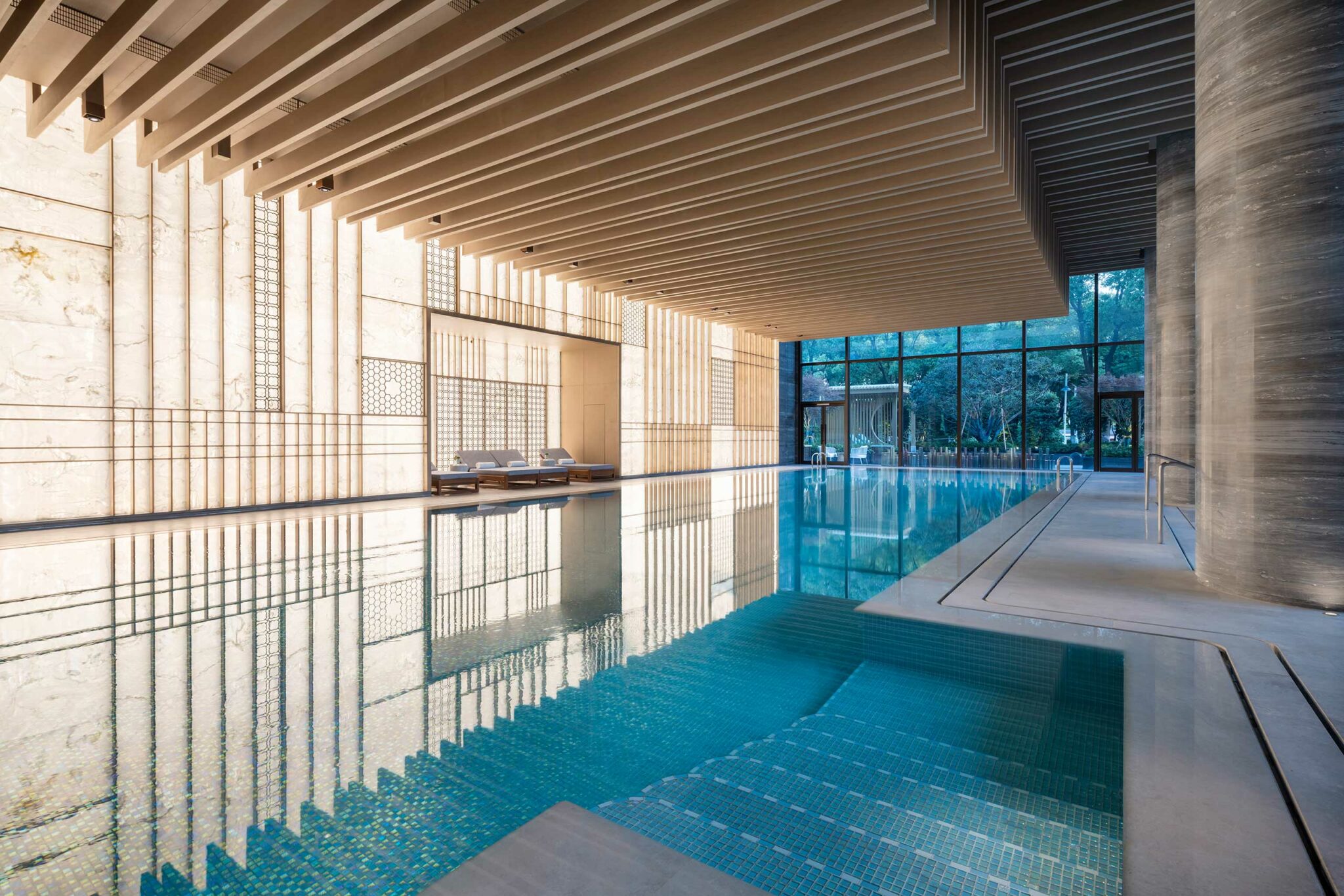 Luxurious indoor swimming pool with modern design, featuring sleek wooden ceilings and ambient lighting. Large windows offer serene views of greenery outside.