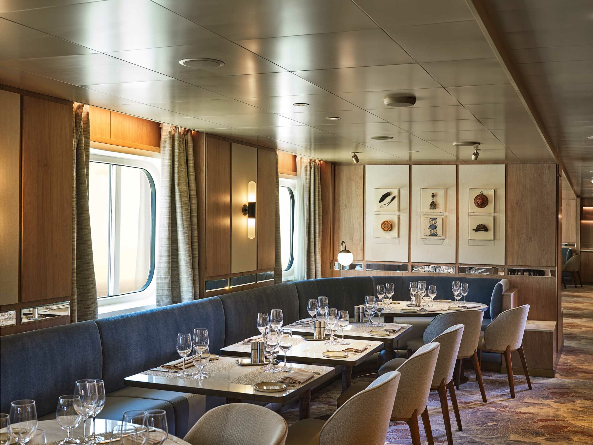 Elegant cruise dining room with soft lighting, featuring neatly set tables, plush seating, large windows, and framed art on wood-paneled walls, creating a cozy atmosphere.