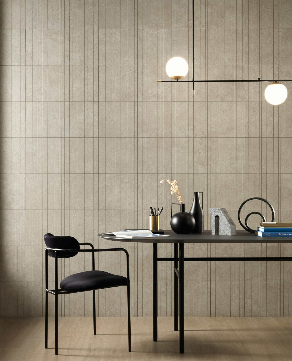 Minimalist workspace with a black chair and table against a textured beige tiled wall. Table holds geometric decor, books, and a suspended globe light. Modern and calm ambiance.