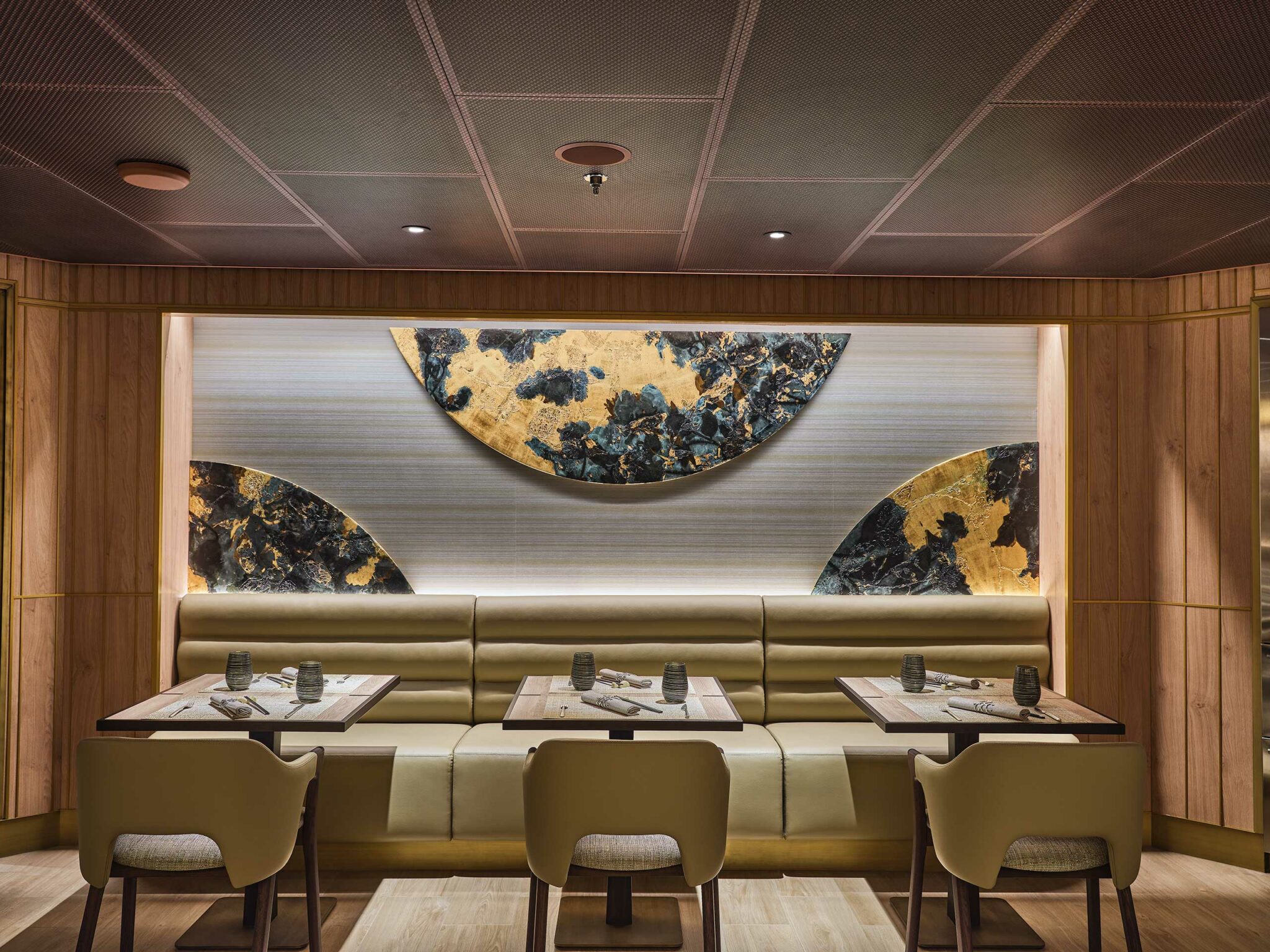Modern cruise restaurant interior with beige seating, wooden tables, and abstract gold and black wall art. Soft lighting creates a cozy, sophisticated ambiance.