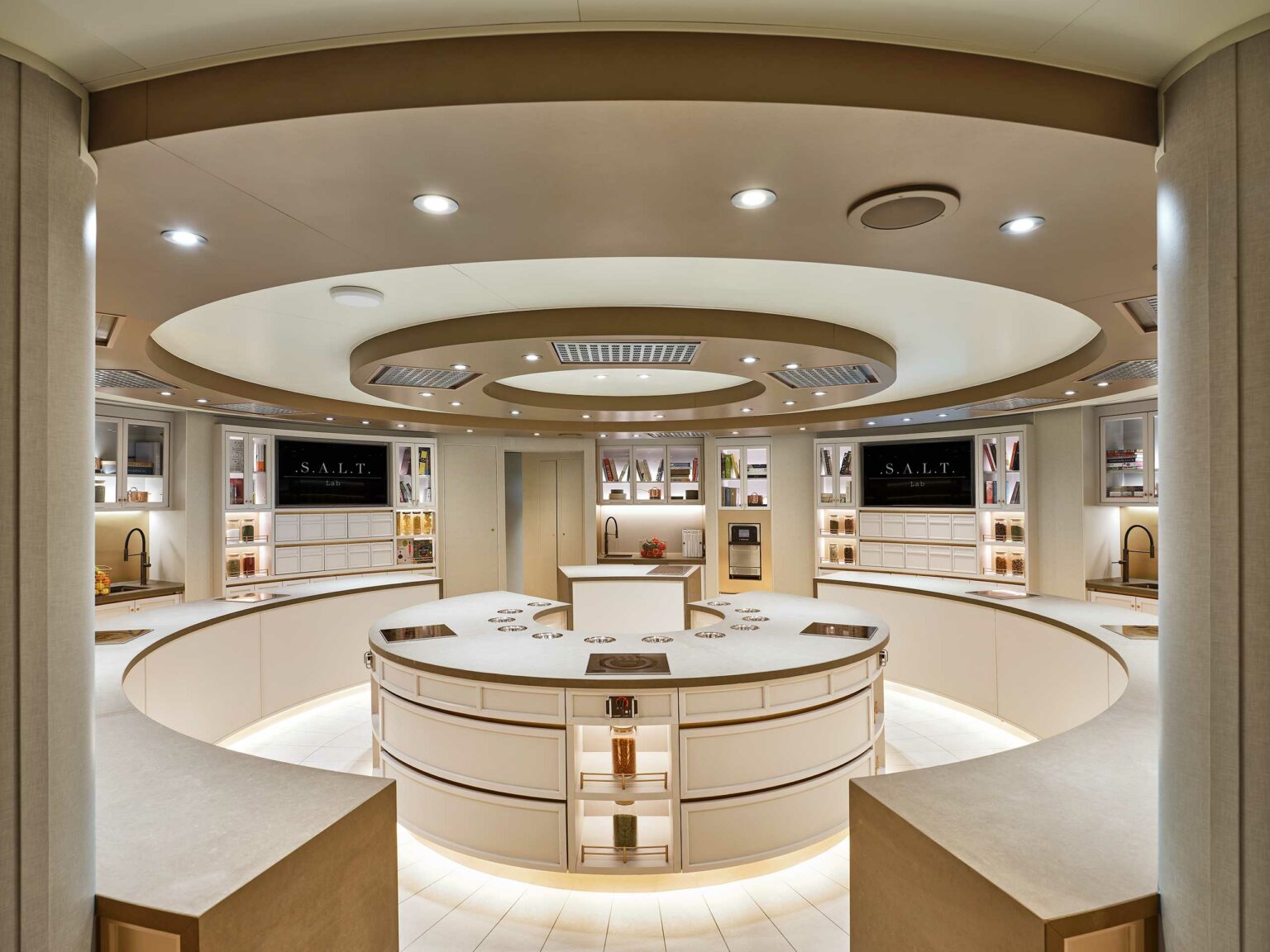 Modern, spacious cruise kitchen with circular islands, sleek white and cream decor, bright lighting, and shelves displaying colorful jars, conveying a clean, sophisticated feel.