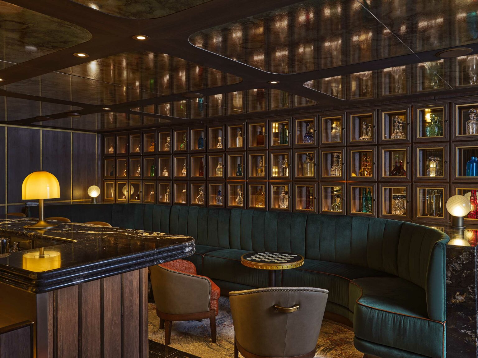 Luxurious cruise bar with a dimly lit ambiance featuring dark wood, a curved velvet sofa, backlit shelves with decorative bottles, and small round tables.