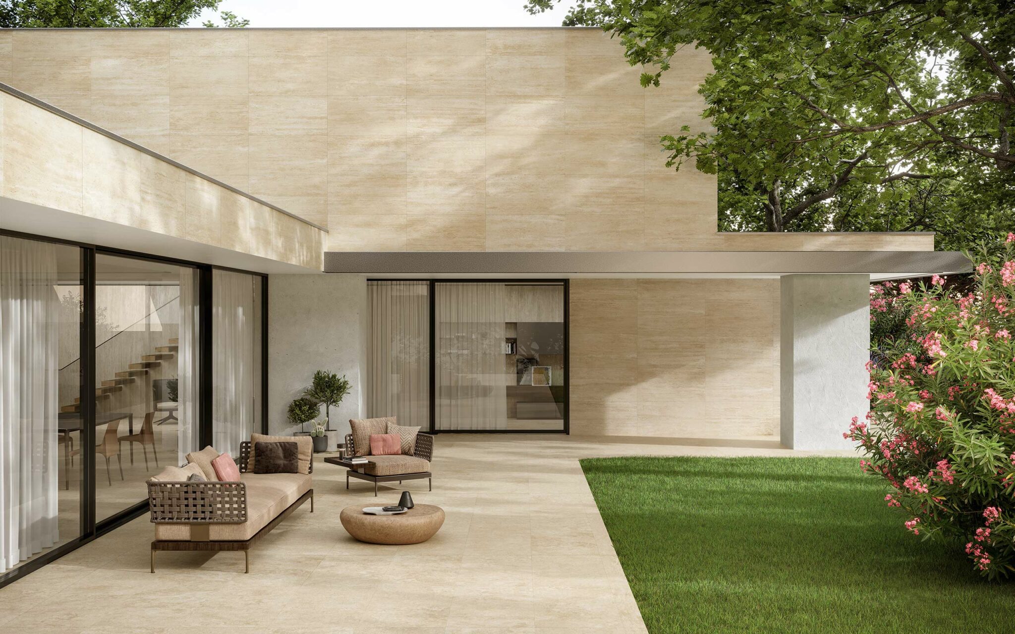 Modern patio with beige stone flooring, wicker furniture with cushions, glass sliding doors, green lawn, and blooming pink flowers. Calm, inviting atmosphere.