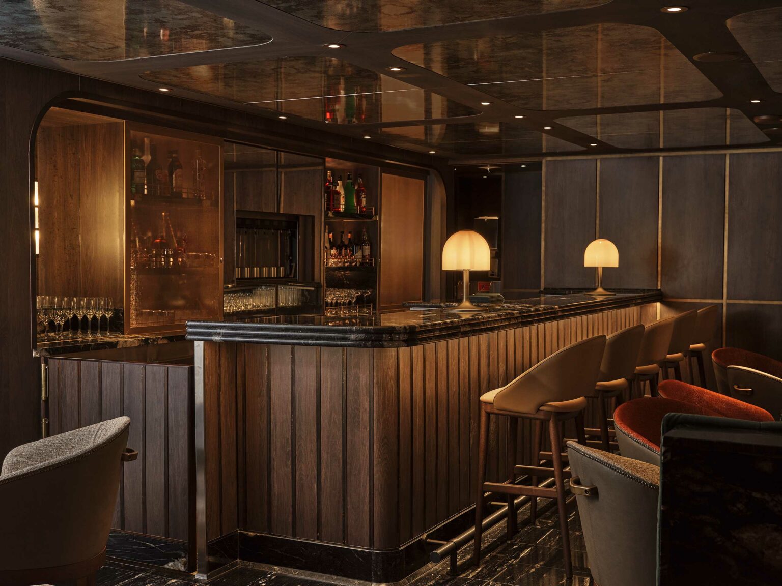 Sophisticated cruise bar with wood paneling, black marble countertop, and soft lighting from two lamps. Stools are aligned along the bar, creating an inviting ambiance.