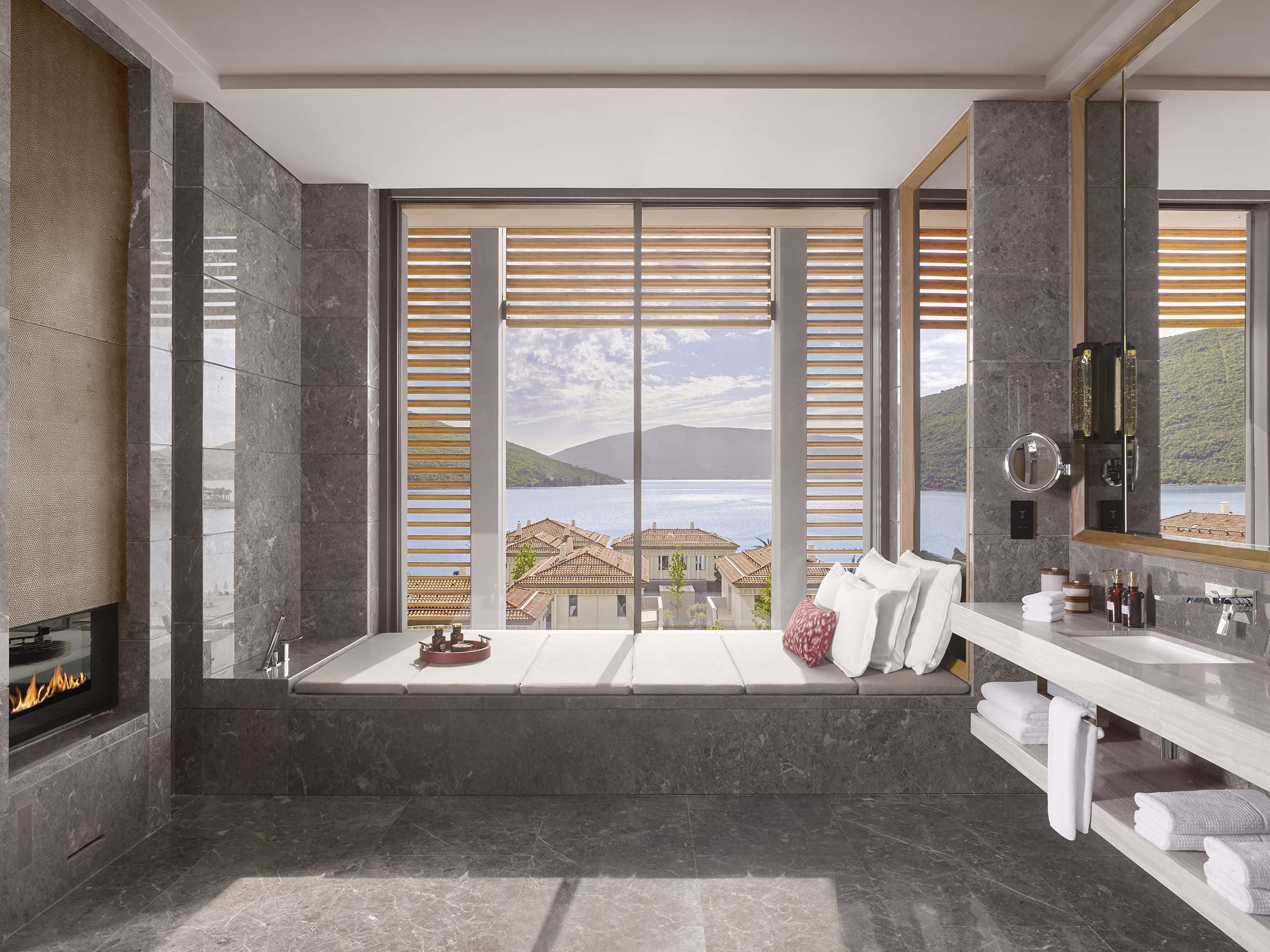 Luxurious bathroom with a large window overlooking a serene sea view and hills. Features elegant stone tiles, a bathtub, fireplace, and modern fixtures.