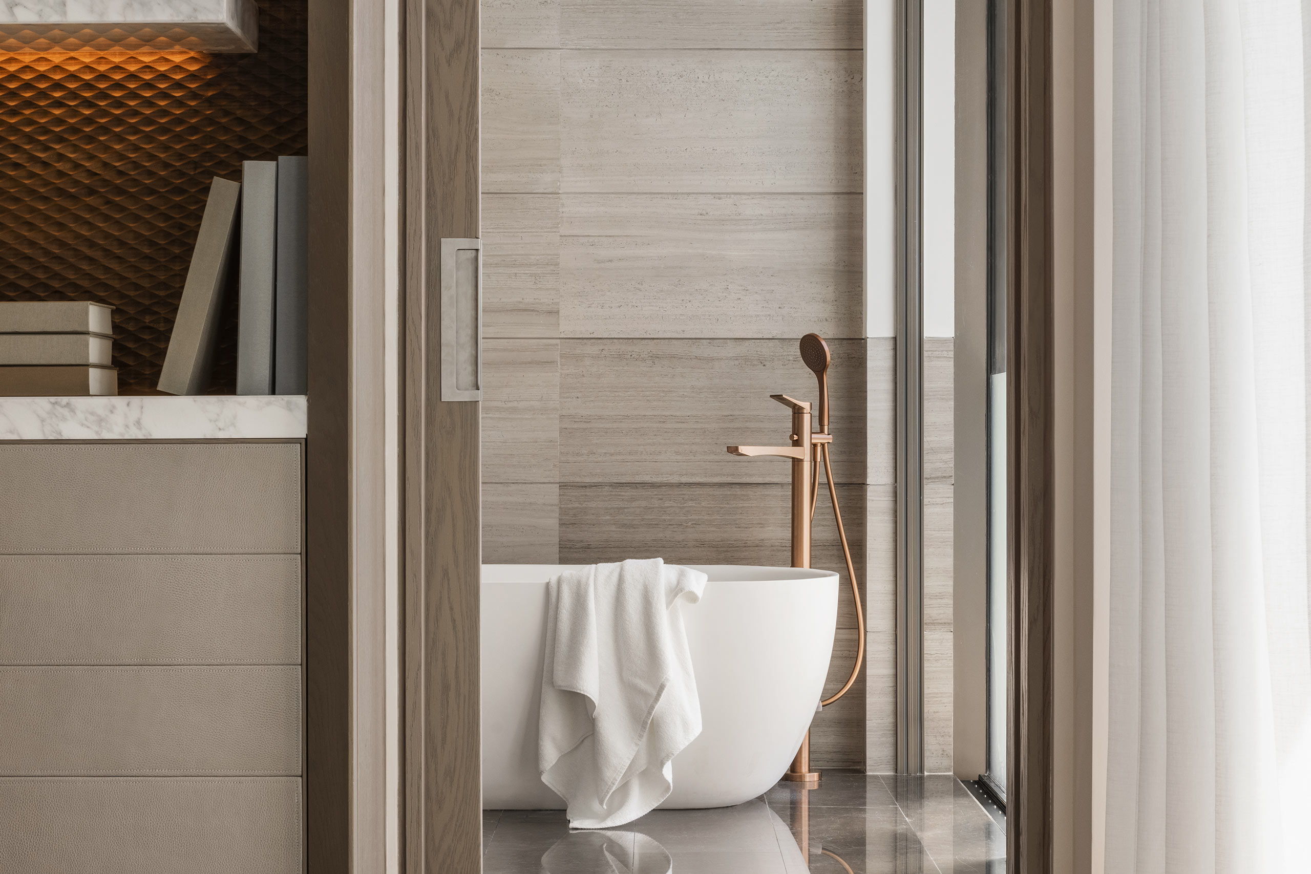 Modern bathroom with a freestanding white tub and copper faucet against textured beige tiles. A soft white towel hangs over the tub, conveying a serene atmosphere.