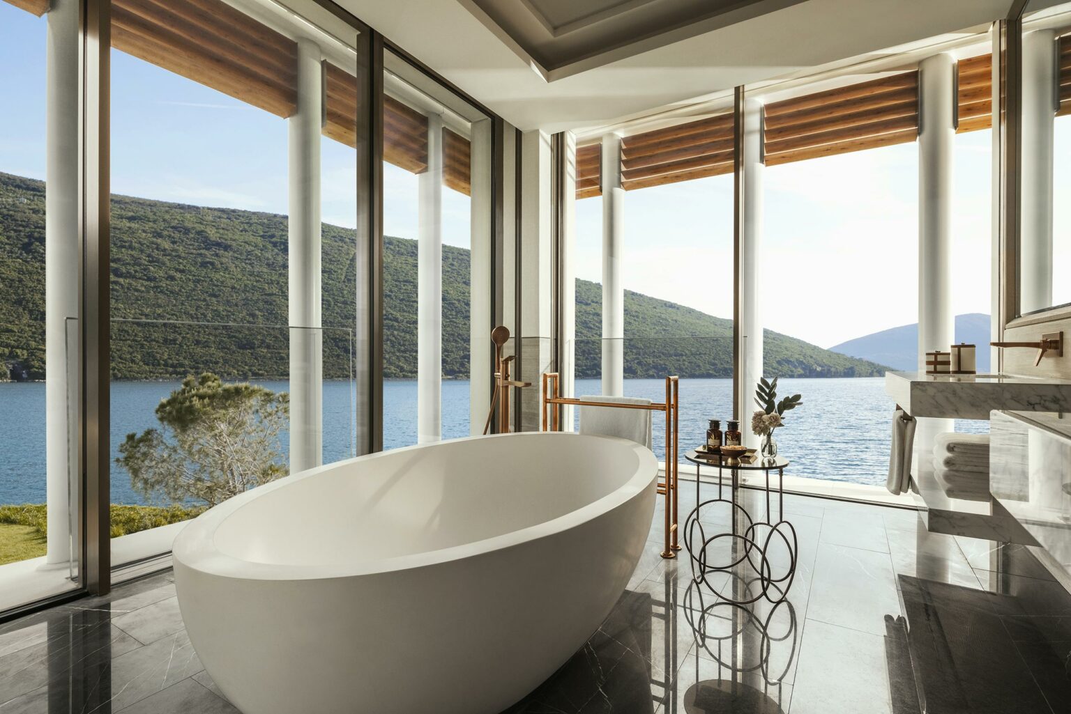 Luxurious bathroom with an oval tub overlooking a scenic lake and green hills through tall glass windows. Elegant, serene, and modern atmosphere.