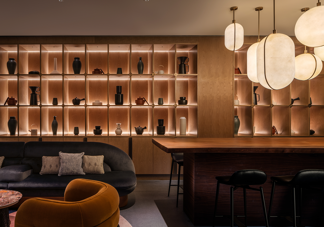 Modern lounge with warm lighting showcases wooden shelves filled with elegant vases and teapots. Cozy seating, dark sofa, and stylish pendant lights.