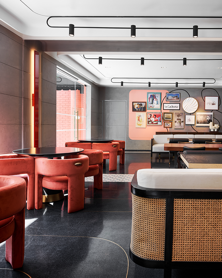 Chic restaurant interior with coral velvet chairs, black tables, and cane accents. Walls showcase colorful artwork, lit by modern ceiling lights.
