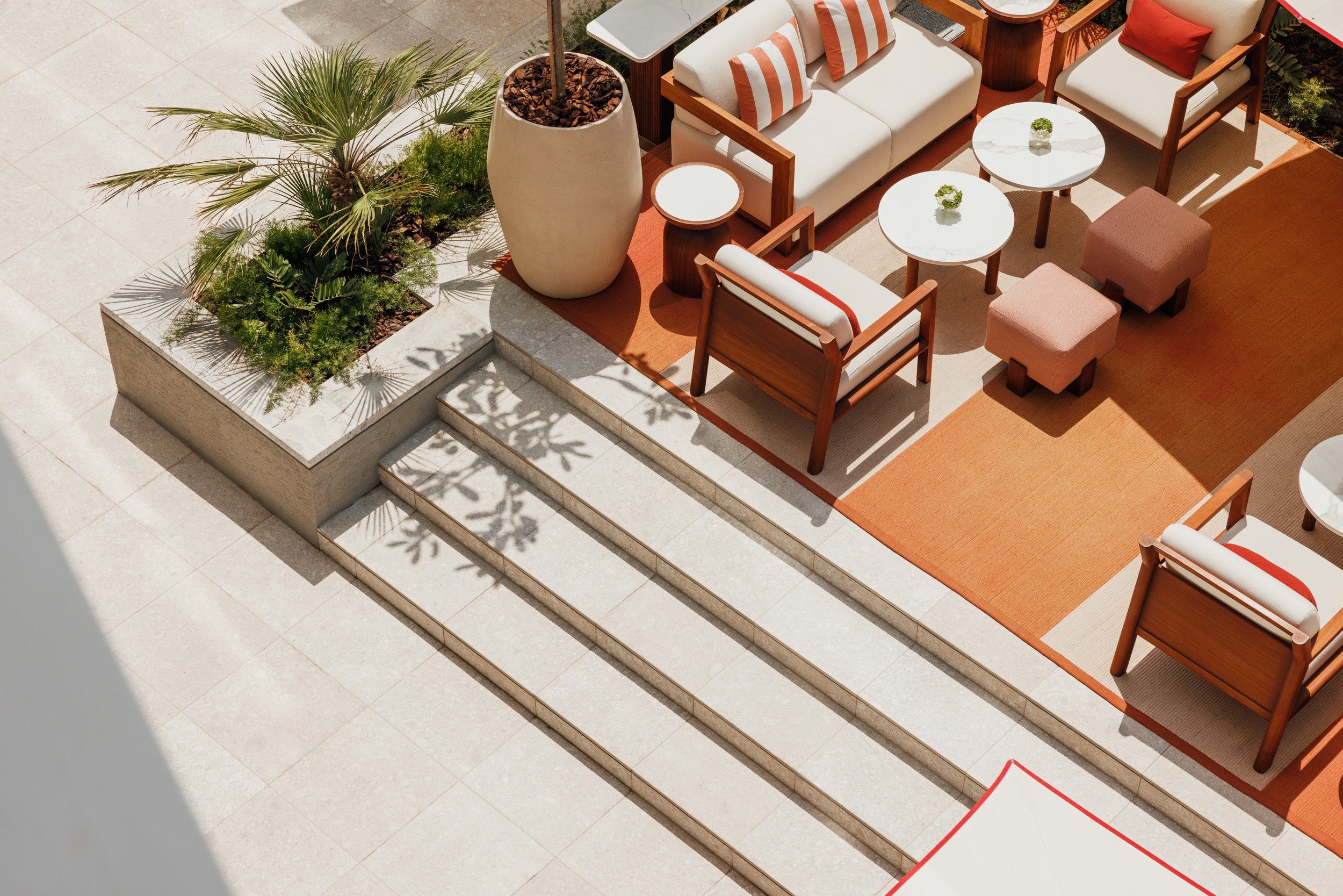 Outdoor seating area with beige and orange furniture, wooden chairs, and small round tables on a sunny patio. Steps lead to a potted plant and greenery.