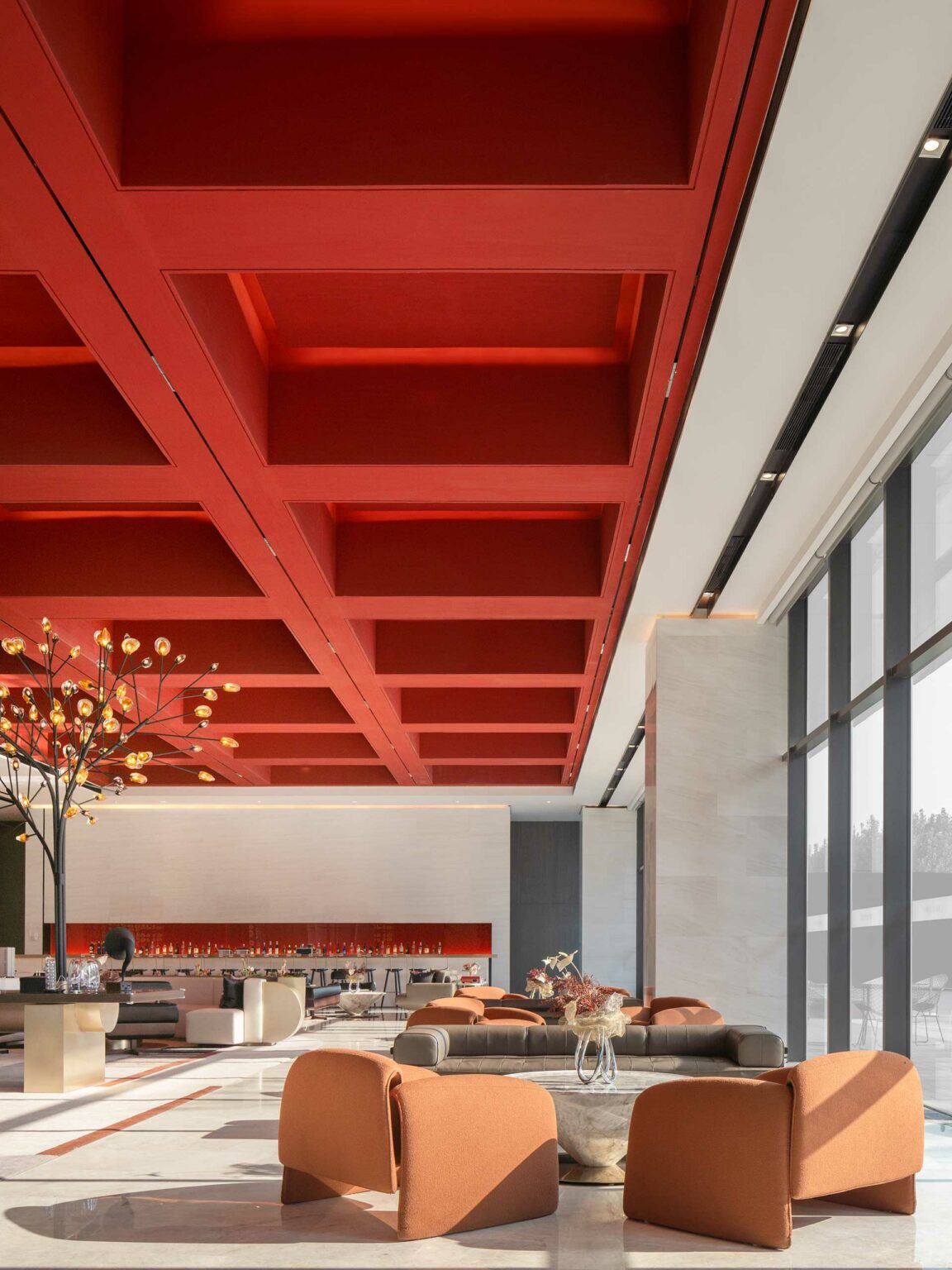 Modern lounge with a red coffered ceiling, featuring expansive windows, stylish seating, and a central decorative tree, creating a welcoming ambiance.