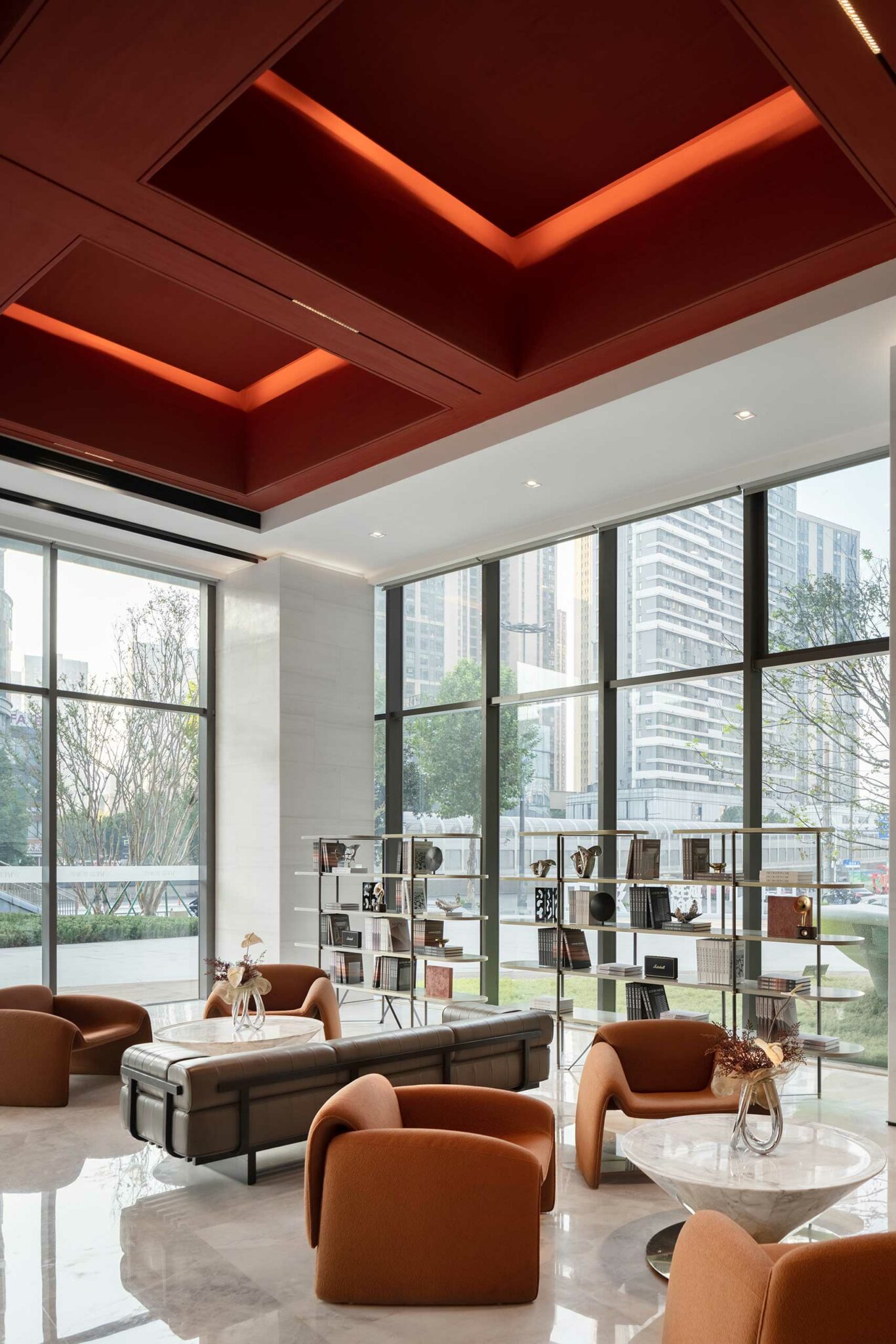 Modern lounge with large windows, brown chairs, and marble tables. A red ceiling adds warmth. Shelves with books and decor create a sophisticated vibe.