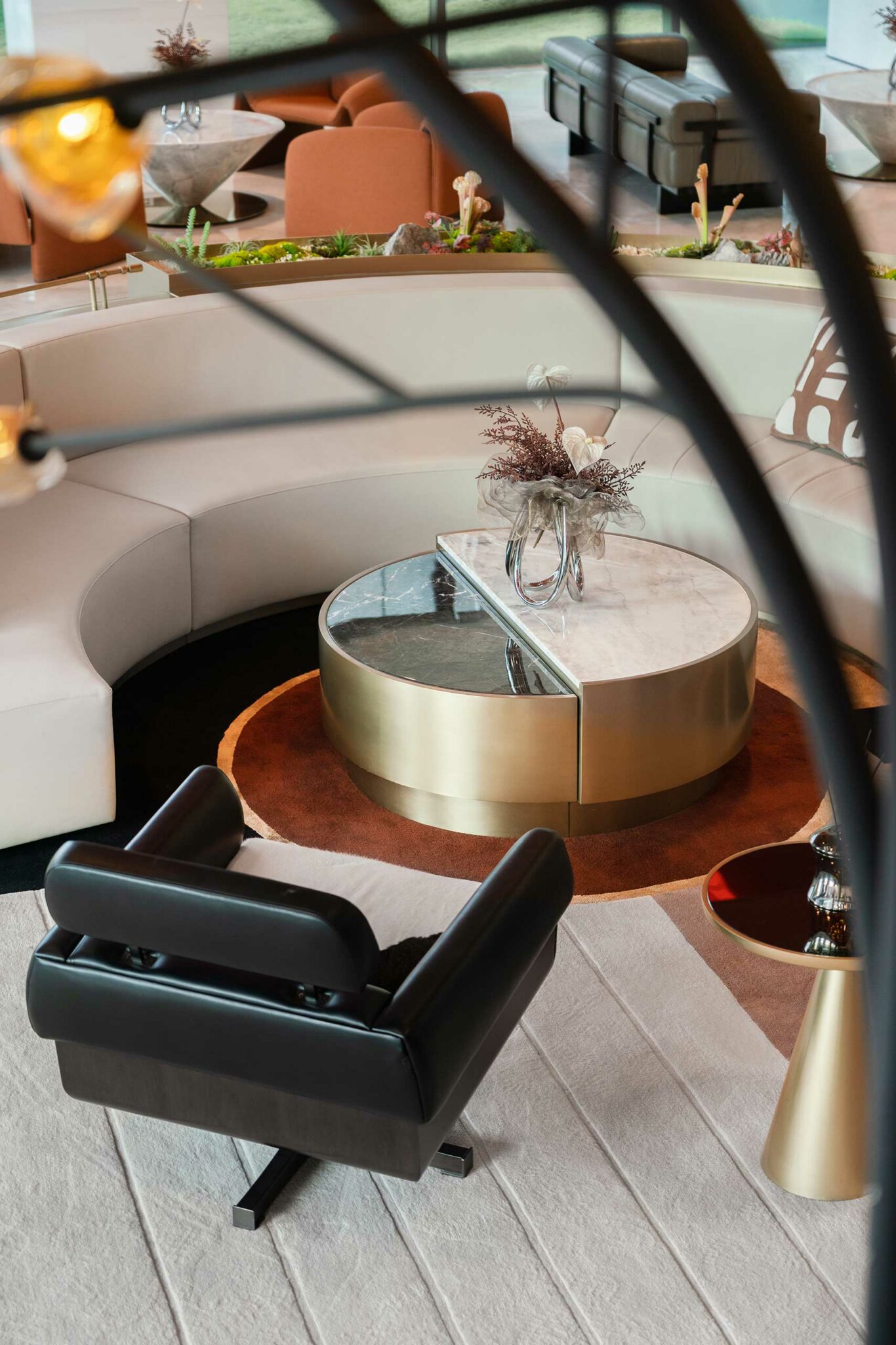 Modern lounge featuring a semi-circular white sofa, central marble and gold table with a glass vase, black chair, and earthy decor. Cozy and elegant.